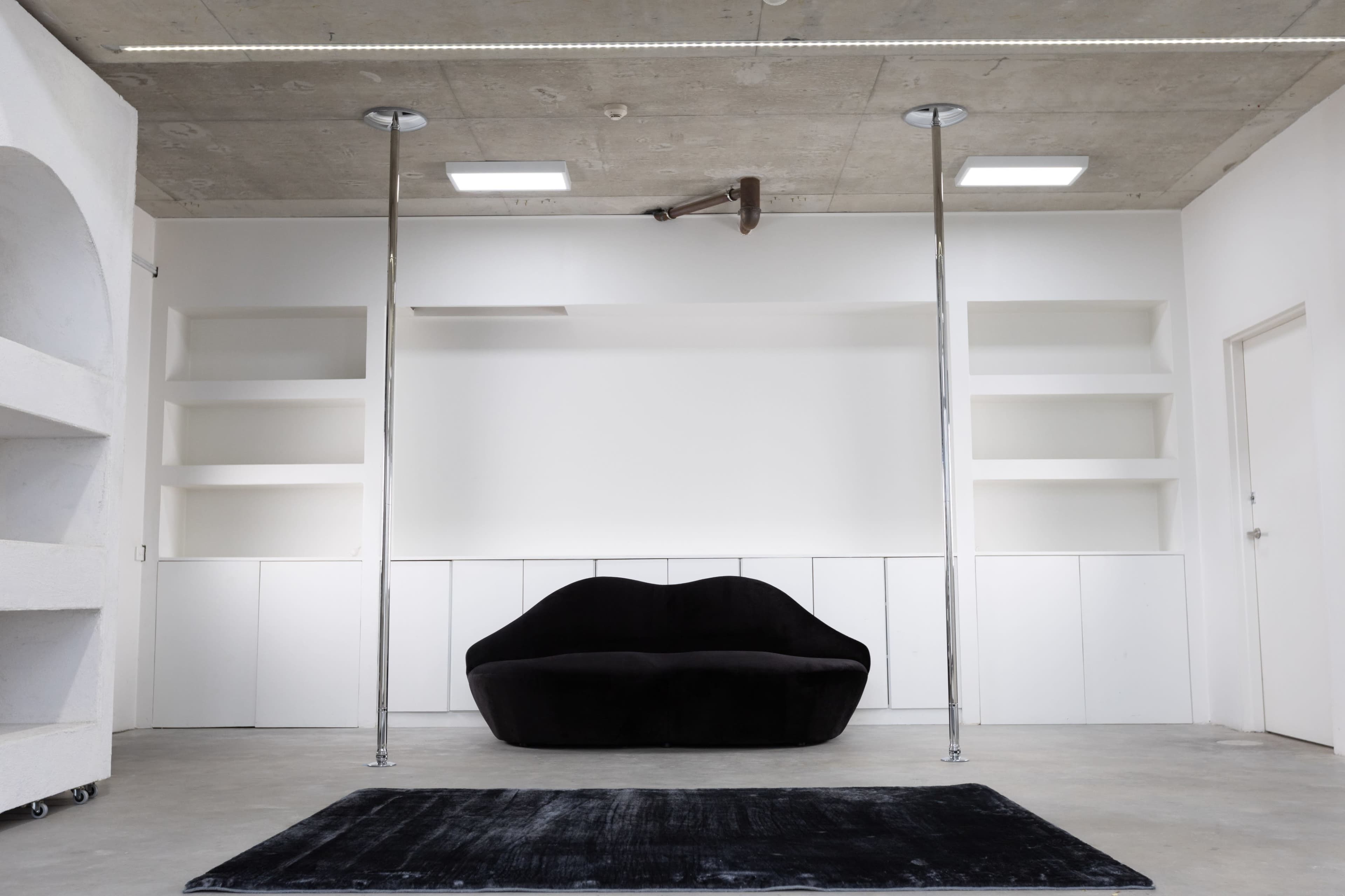A minimalist interior with a black sofa, two standing light fixtures, and white shelves against a concrete wall.