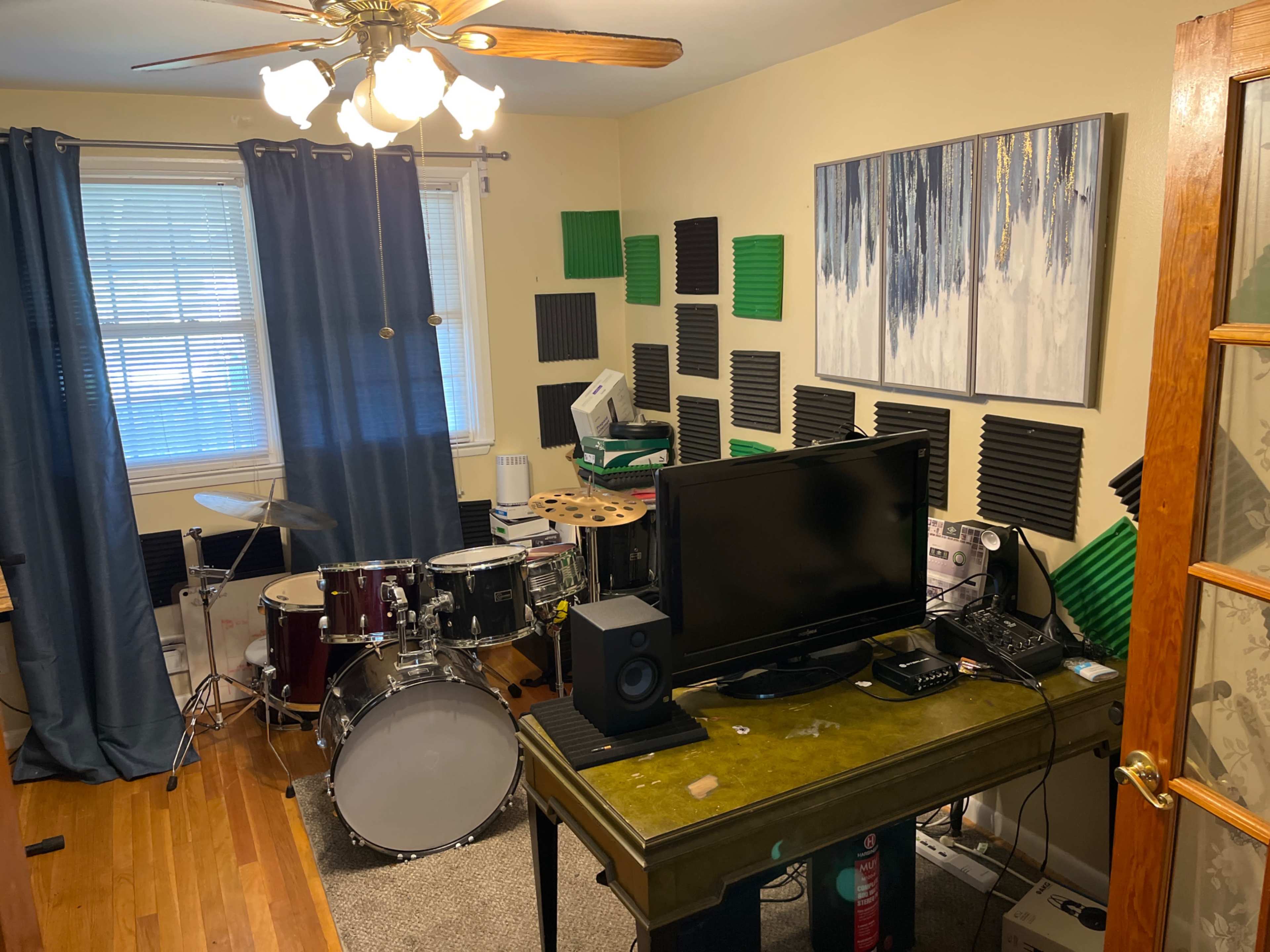 A small room features a drum set, a television, and a desk with electronic equipment, surrounded by soundproofing panels on the walls.