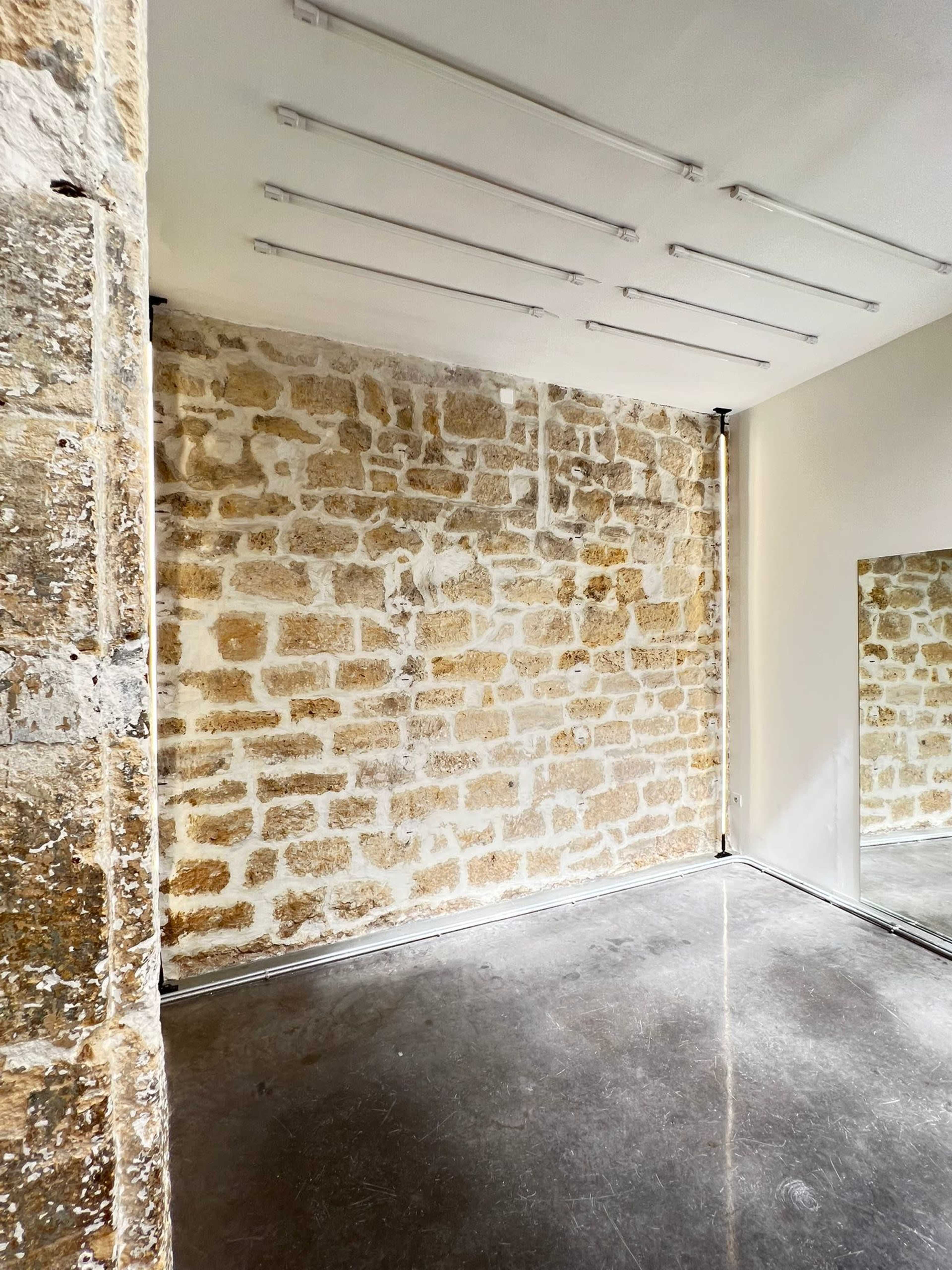 The image shows a minimalist interior space featuring a stone wall, a polished concrete floor, and a large mirror reflecting the room.