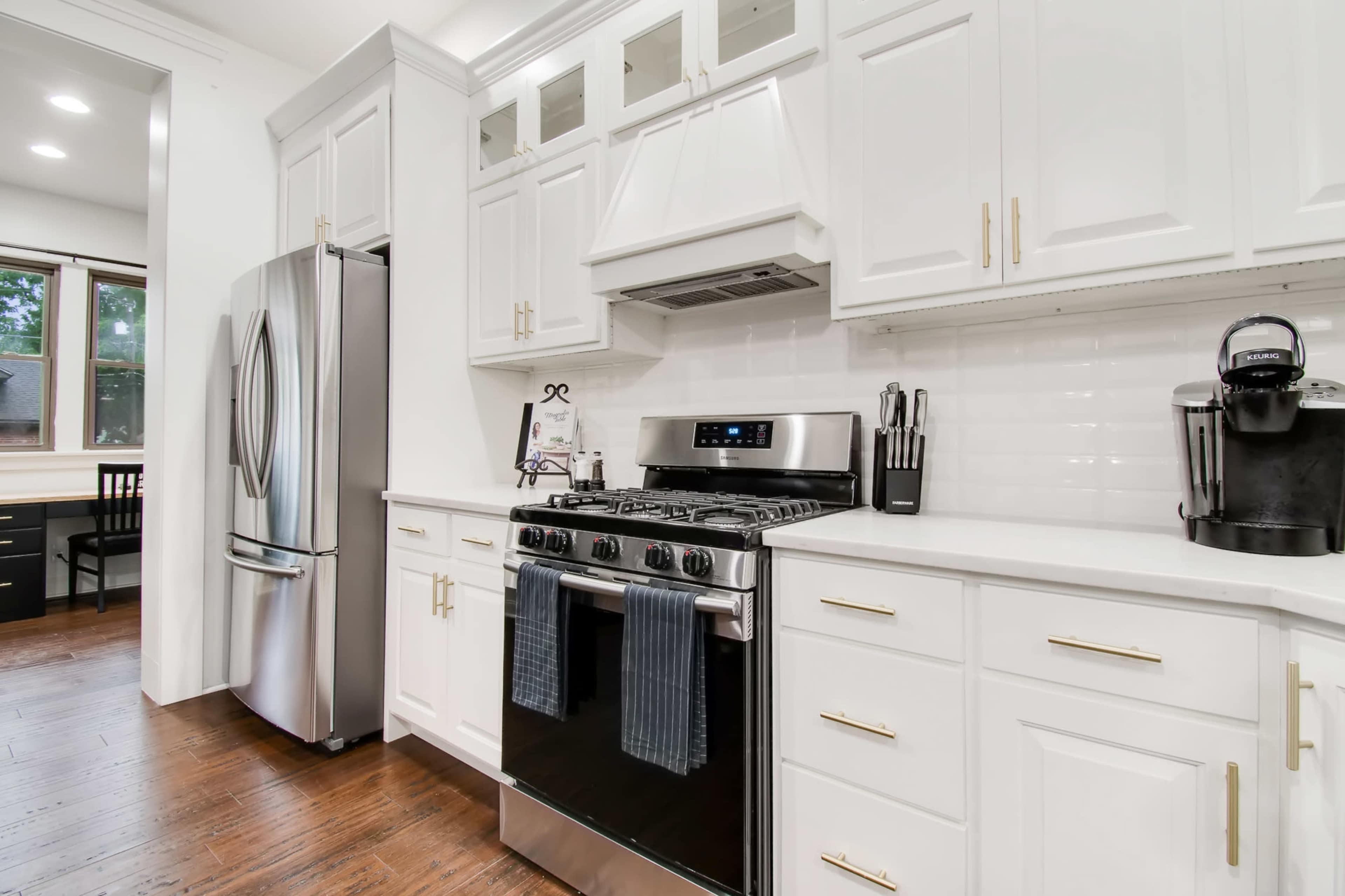 A modern kitchen featuring white cabinetry, a stainless steel refrigerator, a gas stove with a hood, and a dark wood floor.