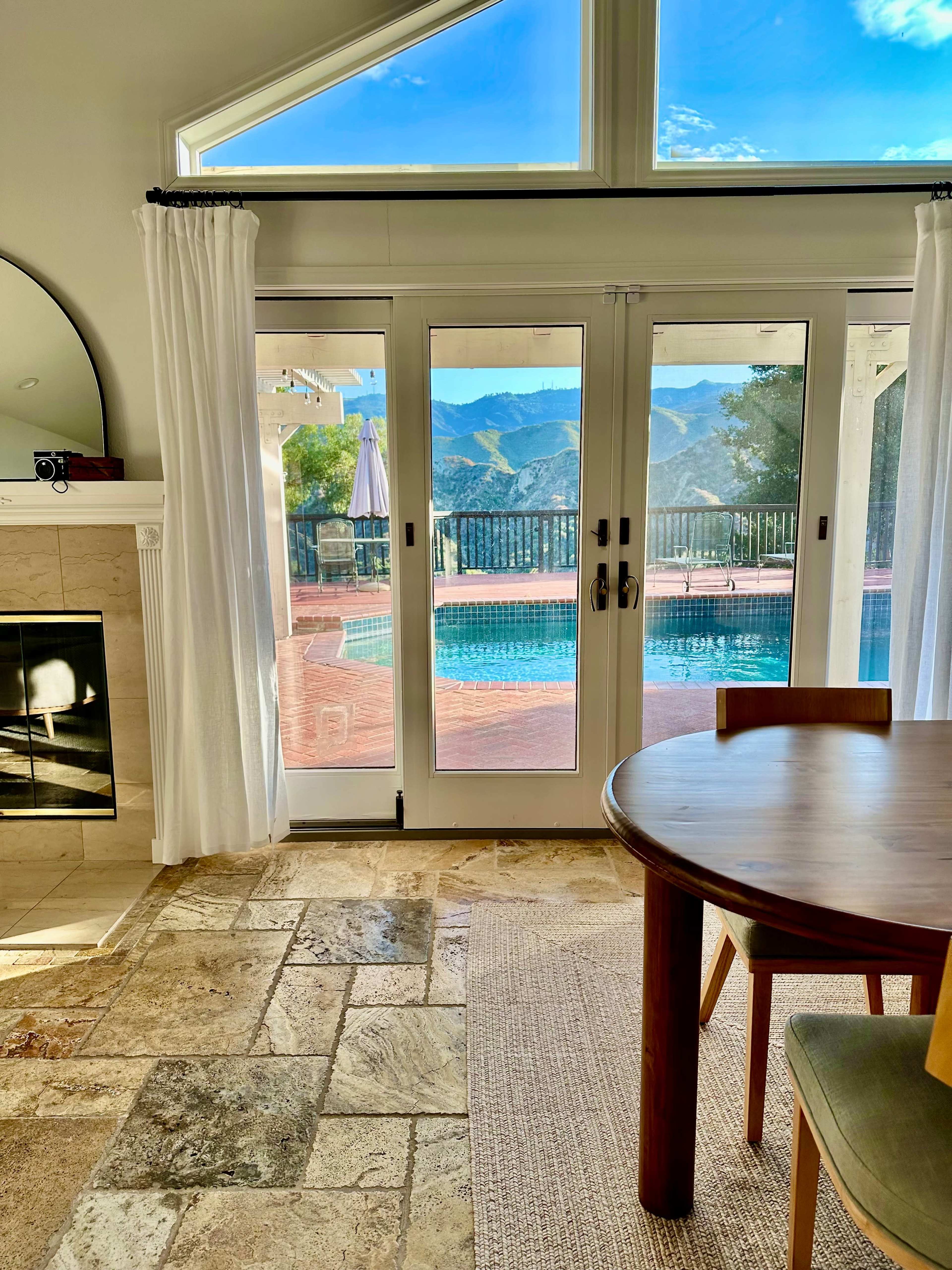 A bright room with a wooden table, a sliding glass door leading to a pool area, and mountains visible in the background.