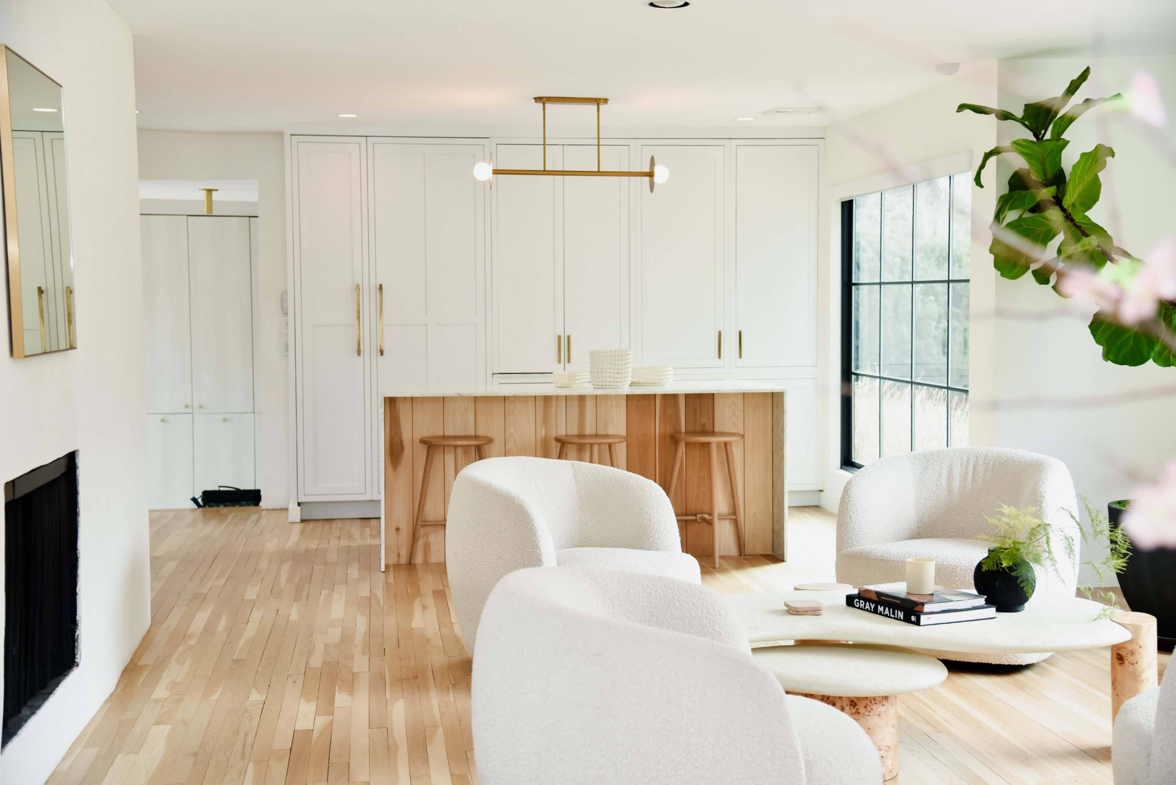 The image shows a modern kitchen and living area featuring light wood flooring, a minimalist kitchen island with bar stools, and large windows allowing natural light.