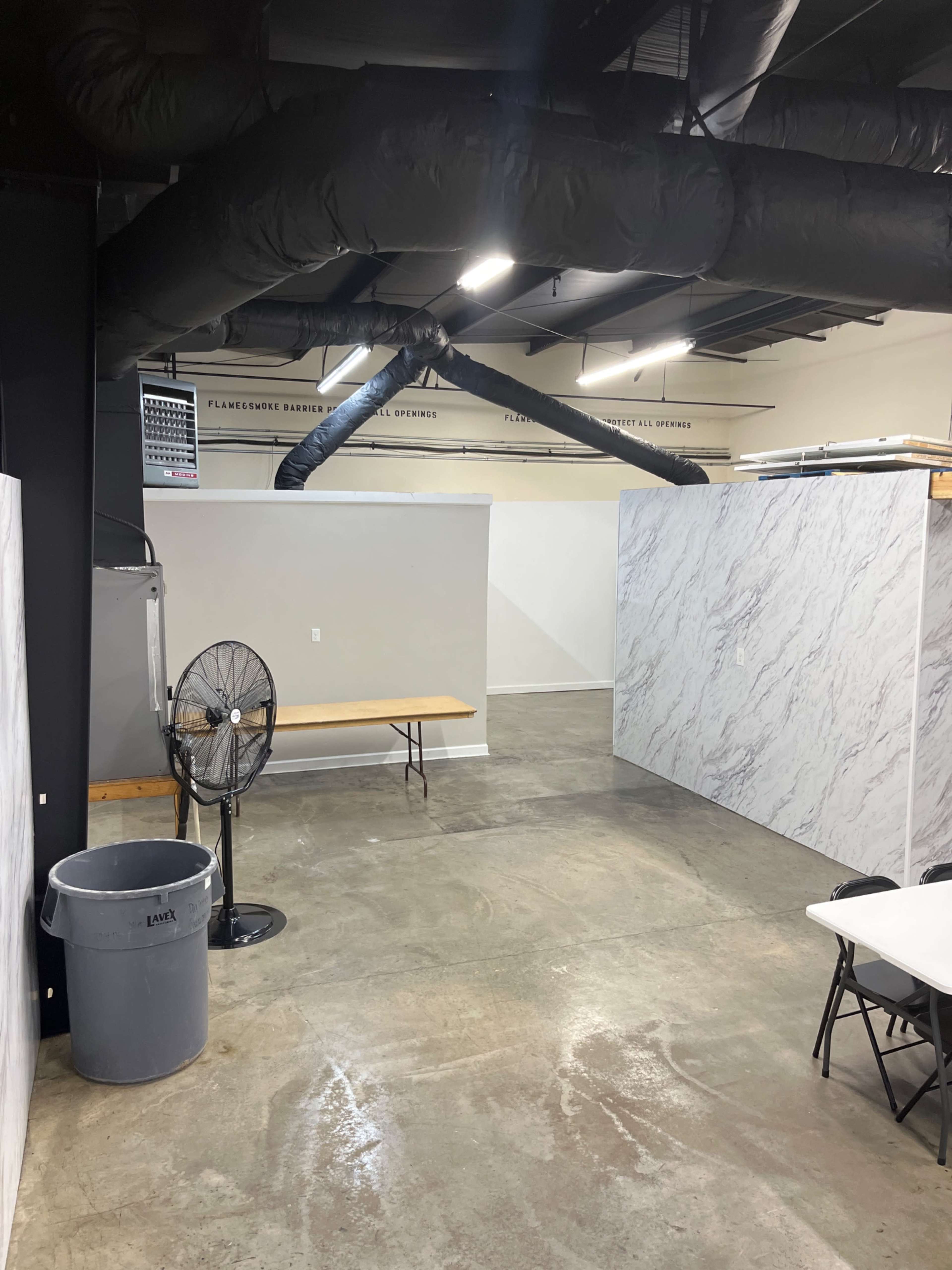 An empty room with concrete flooring, featuring a table along one wall, a fan, and two partition walls.