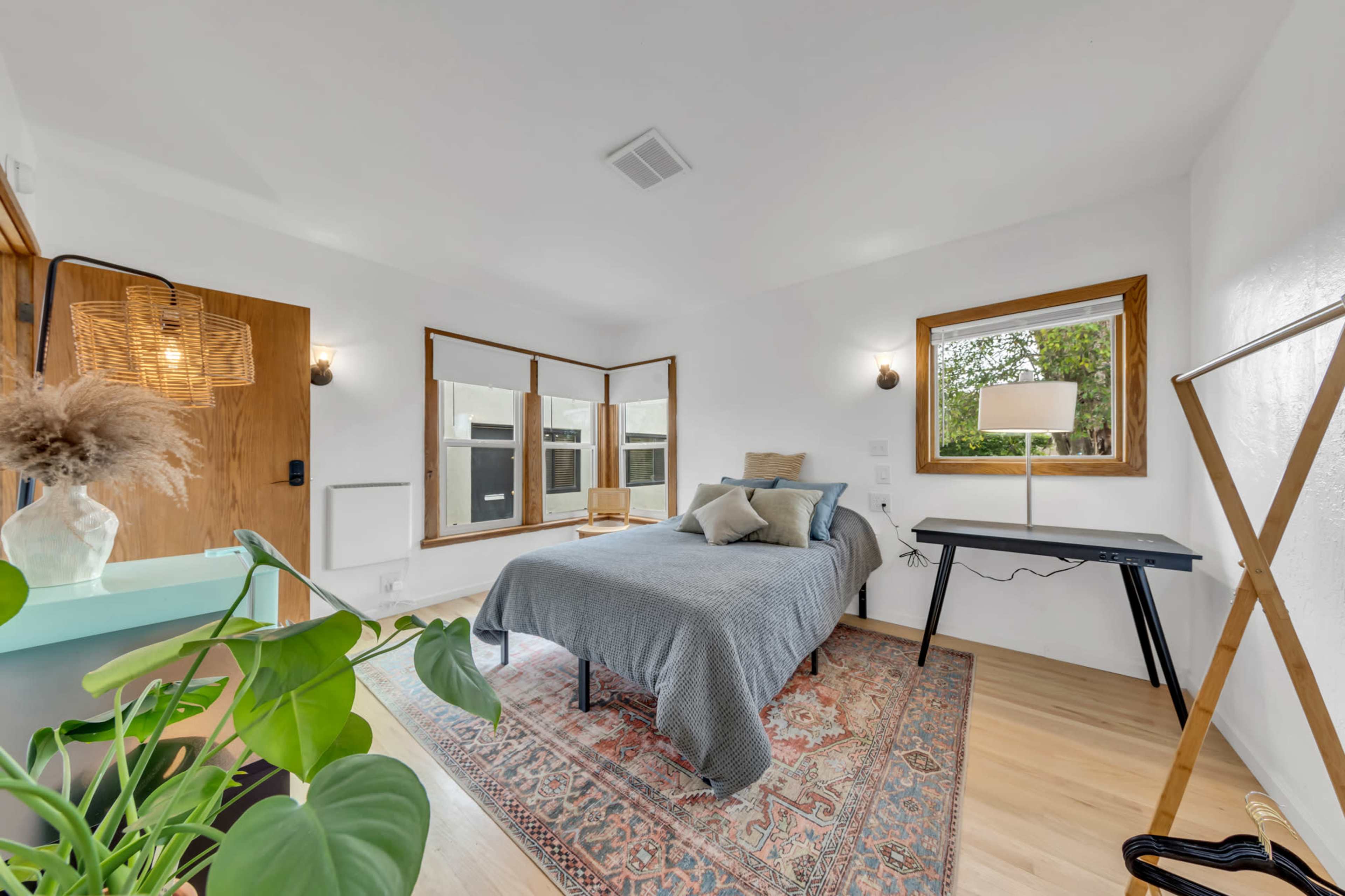 The image shows a neatly arranged bedroom with a bed, a desk, a floor lamp, and a large window, adorned with plants and decorative elements.