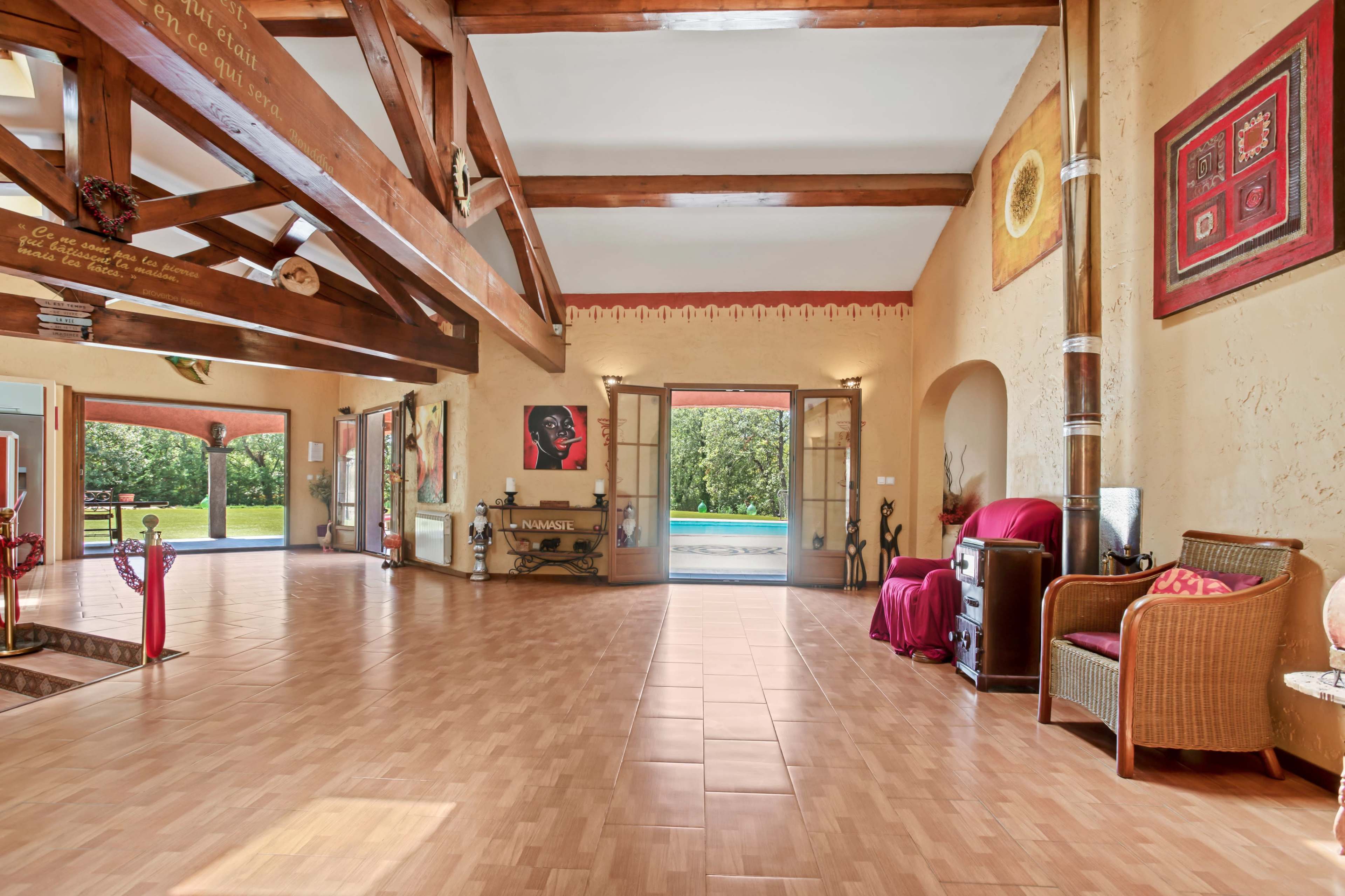 The image shows a spacious room with wooden beams and large doors opening to a garden area, furnished with chairs and decorative elements.