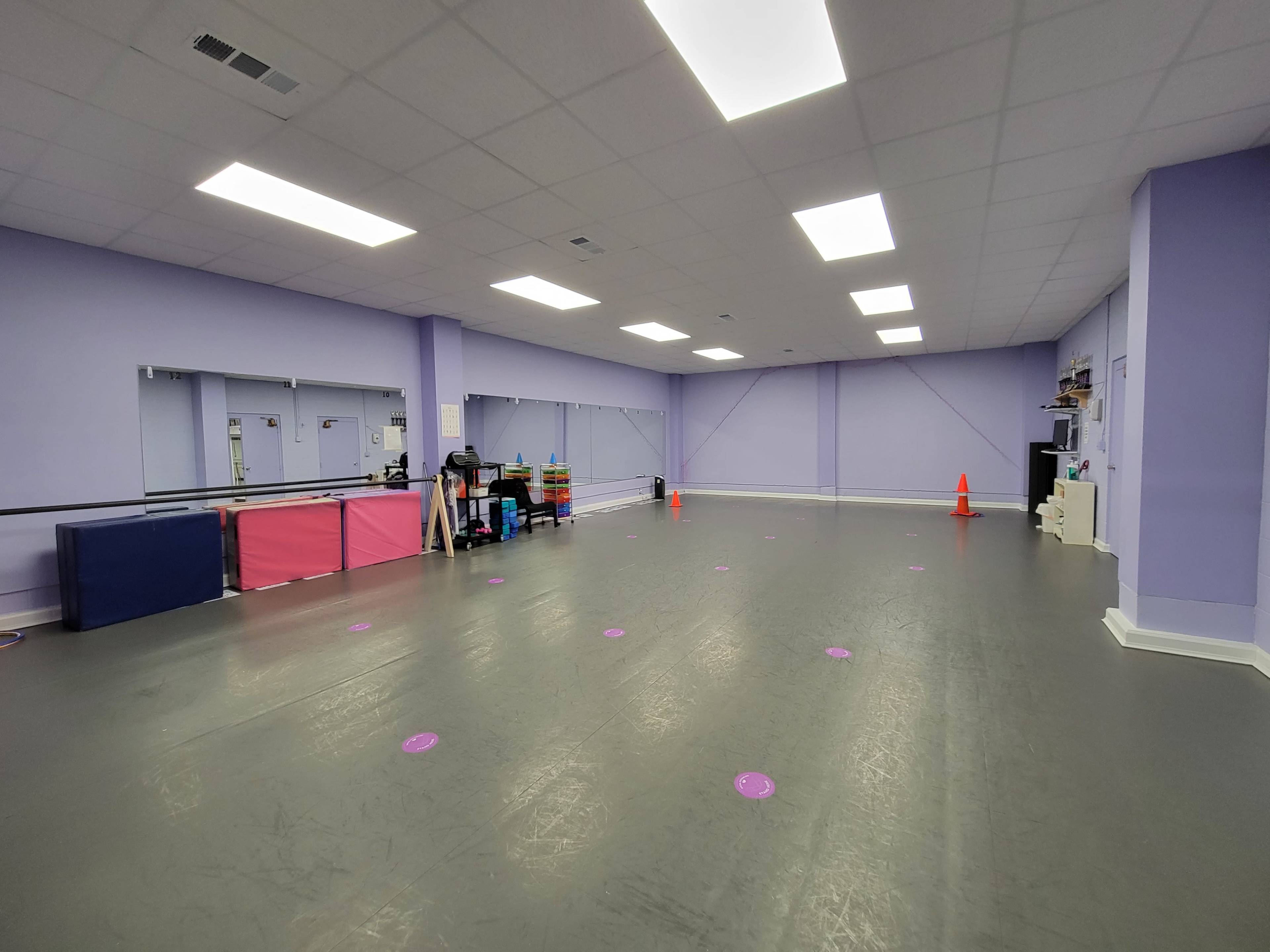 A spacious dance studio with a gray floor, purple walls, and mirrors, featuring some storage equipment and pink mats along one side.
