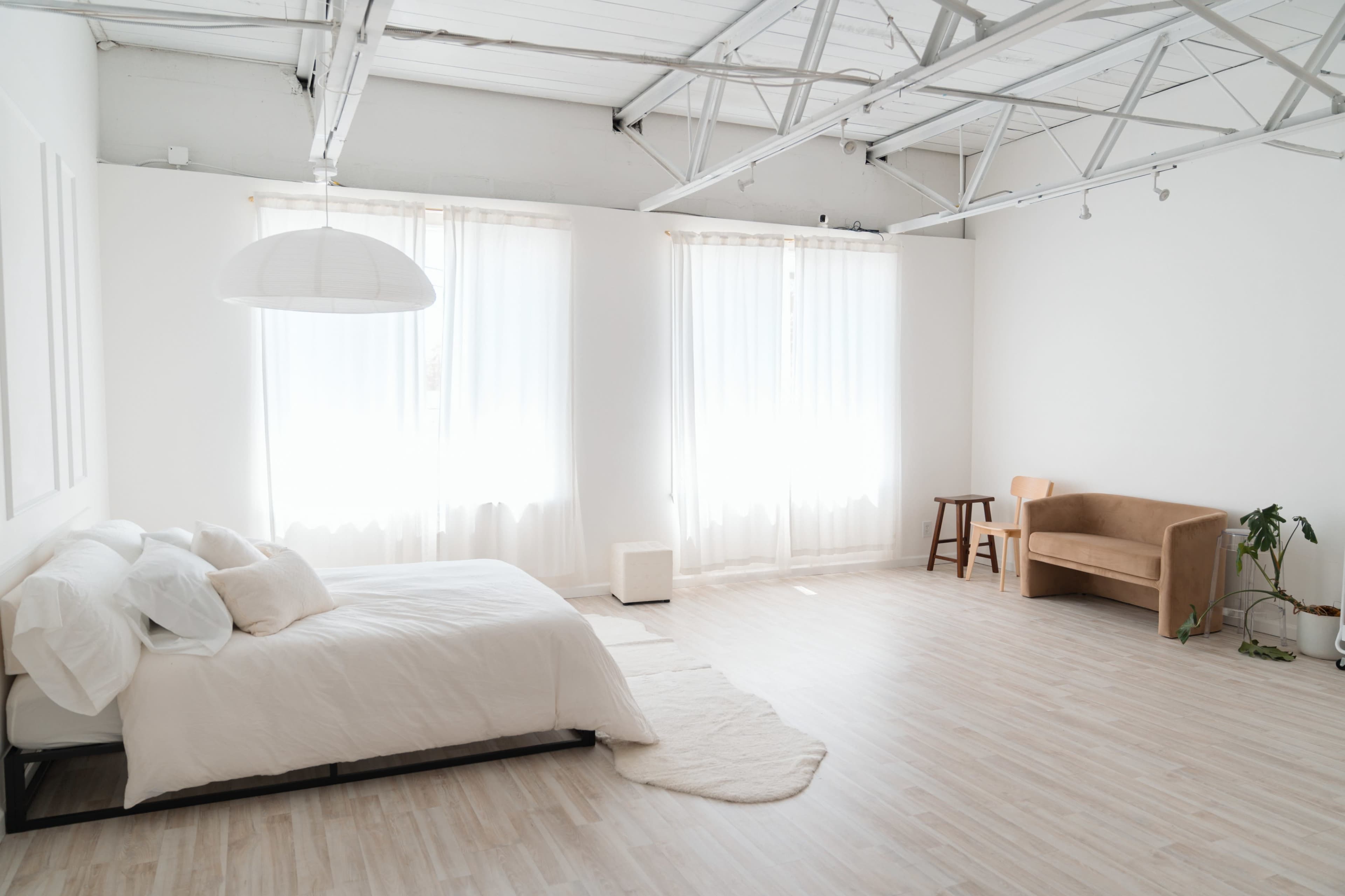 The image shows a minimalist bedroom with a bed, a sofa, and large windows lined with sheer curtains.