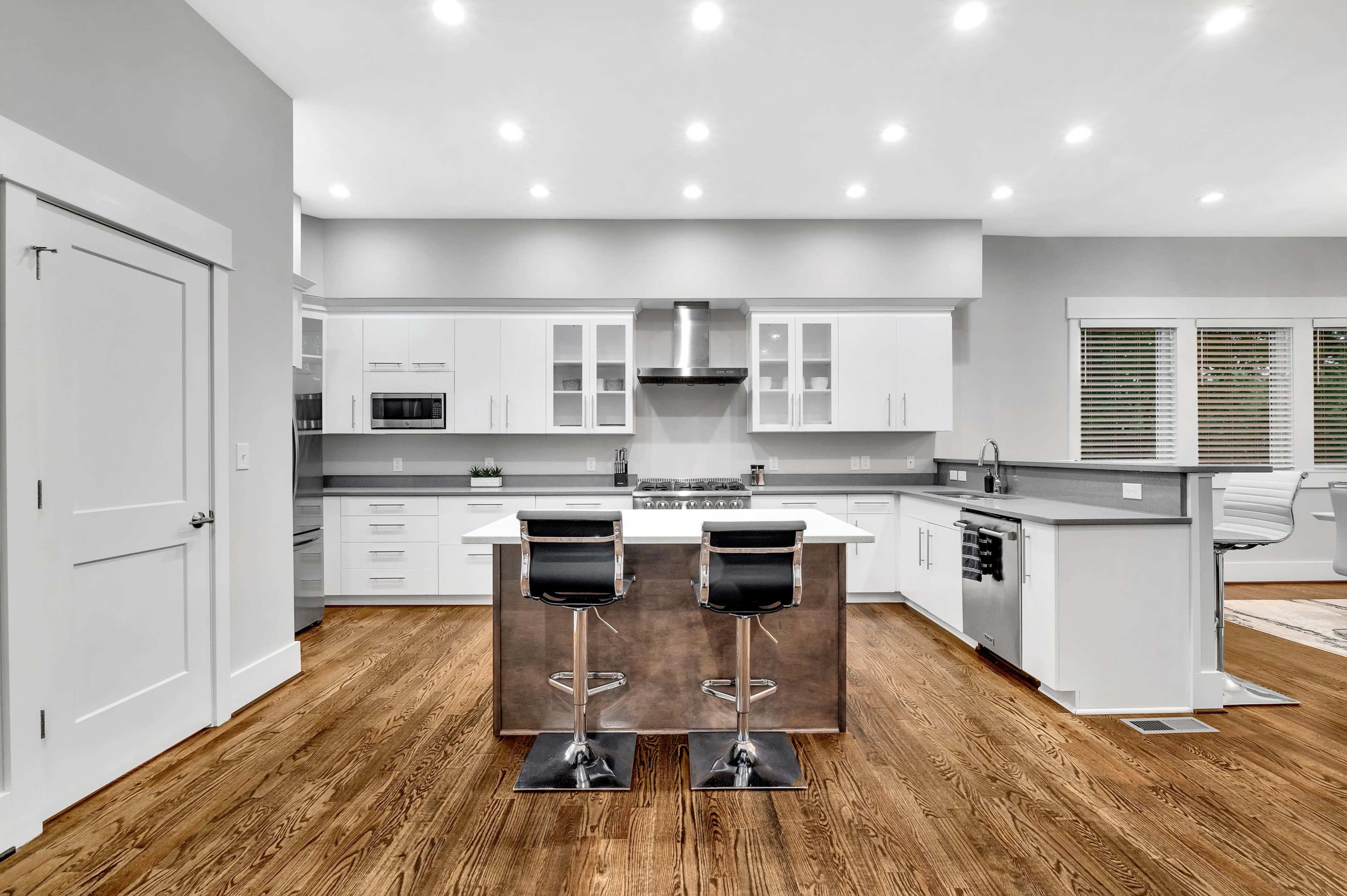 A modern kitchen features white cabinets, a large island with two bar stools, stainless steel appliances, and hardwood flooring.