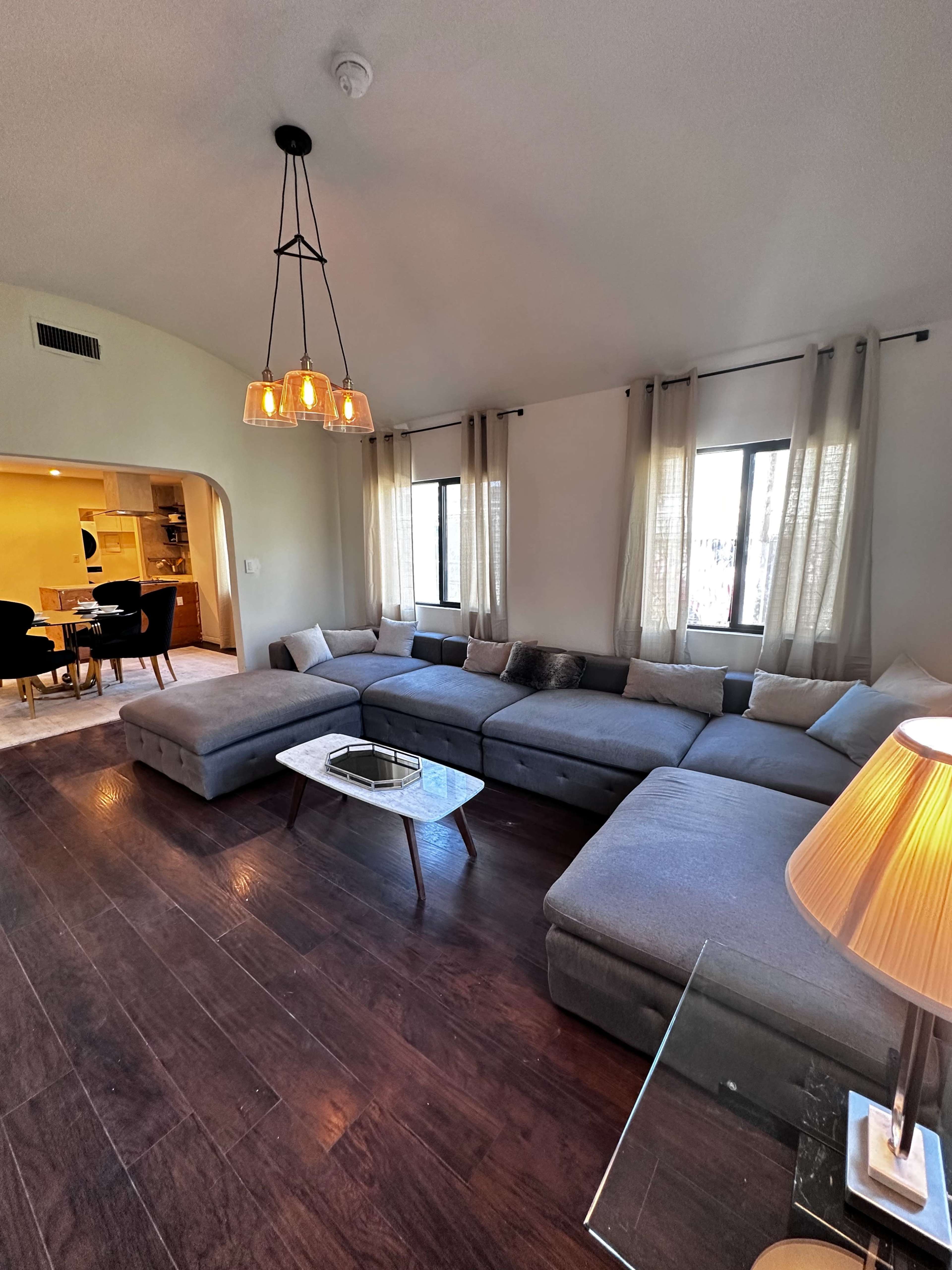 The image shows a modern living room with a large sectional sofa, a coffee table, and windows dressed with curtains, alongside a dining area visible in the background.