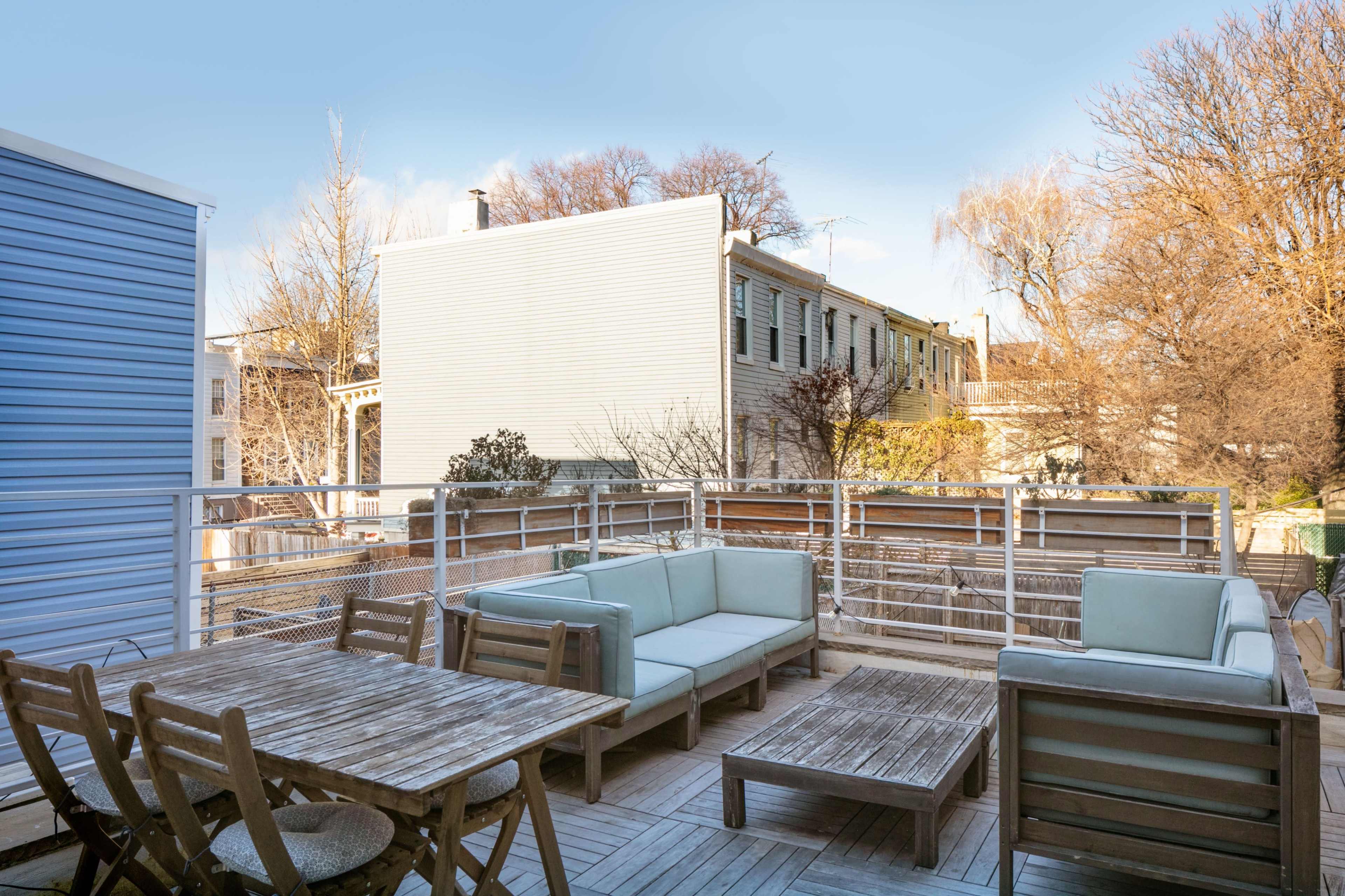 A spacious wooden deck features outdoor furniture and overlooks neighboring buildings under a clear blue sky.