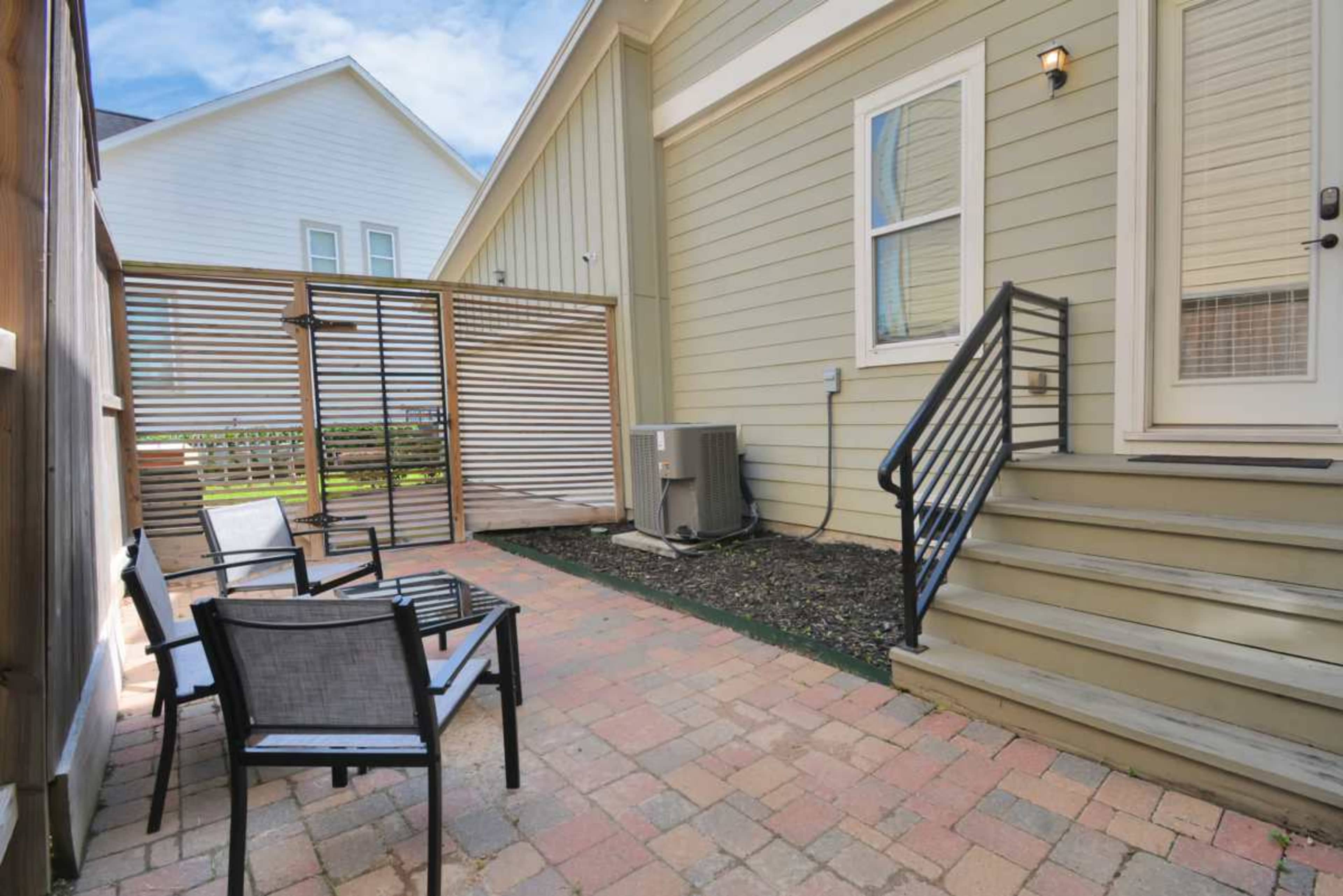 The image shows a small patio area with brick flooring, featuring four chairs, a table, and a lattice fence, alongside a building.