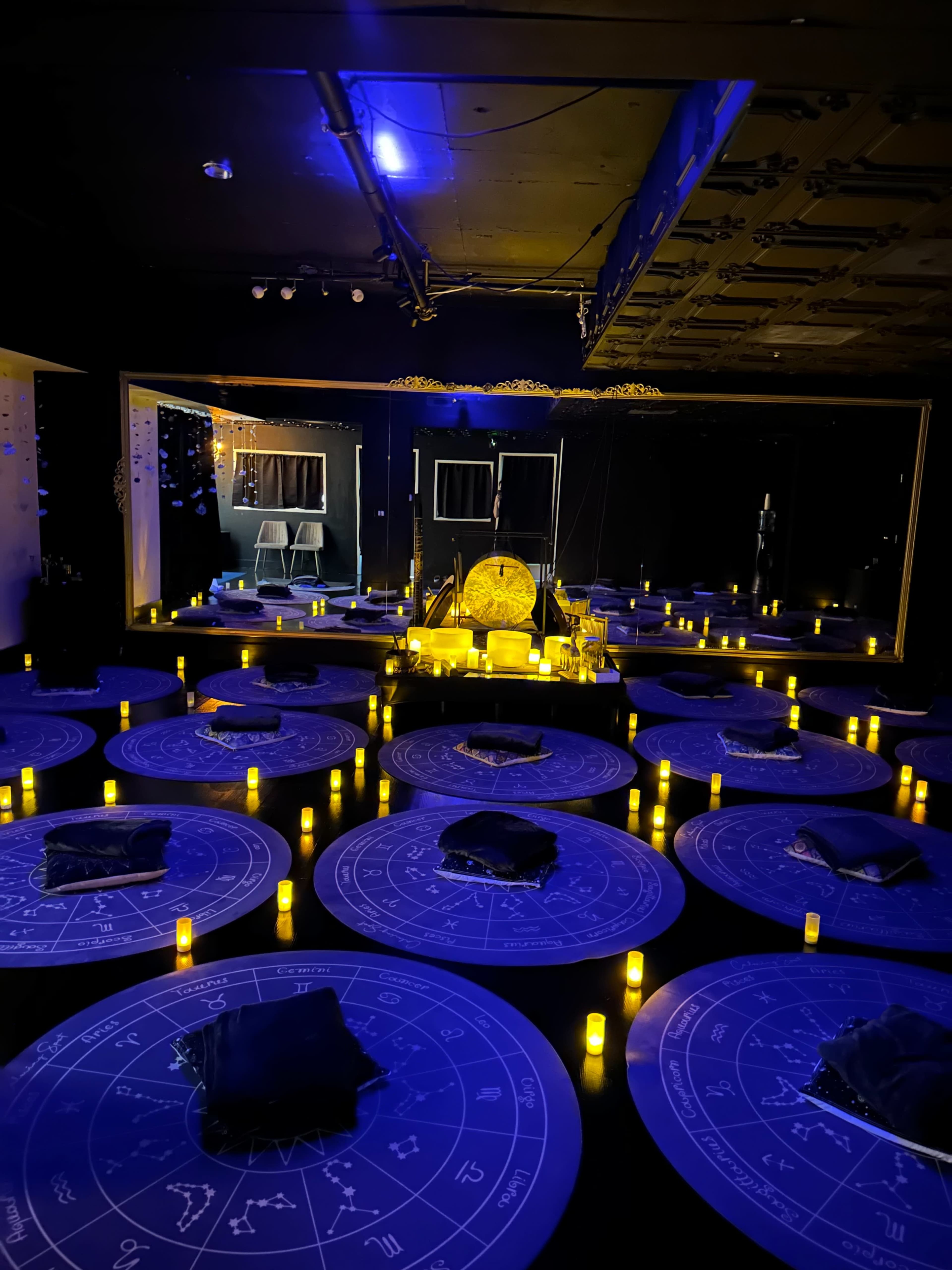 A dimly lit room decorated with circular tables, each featuring astrological symbols, surrounded by flickering candles and a central illuminated area with an ornate object.