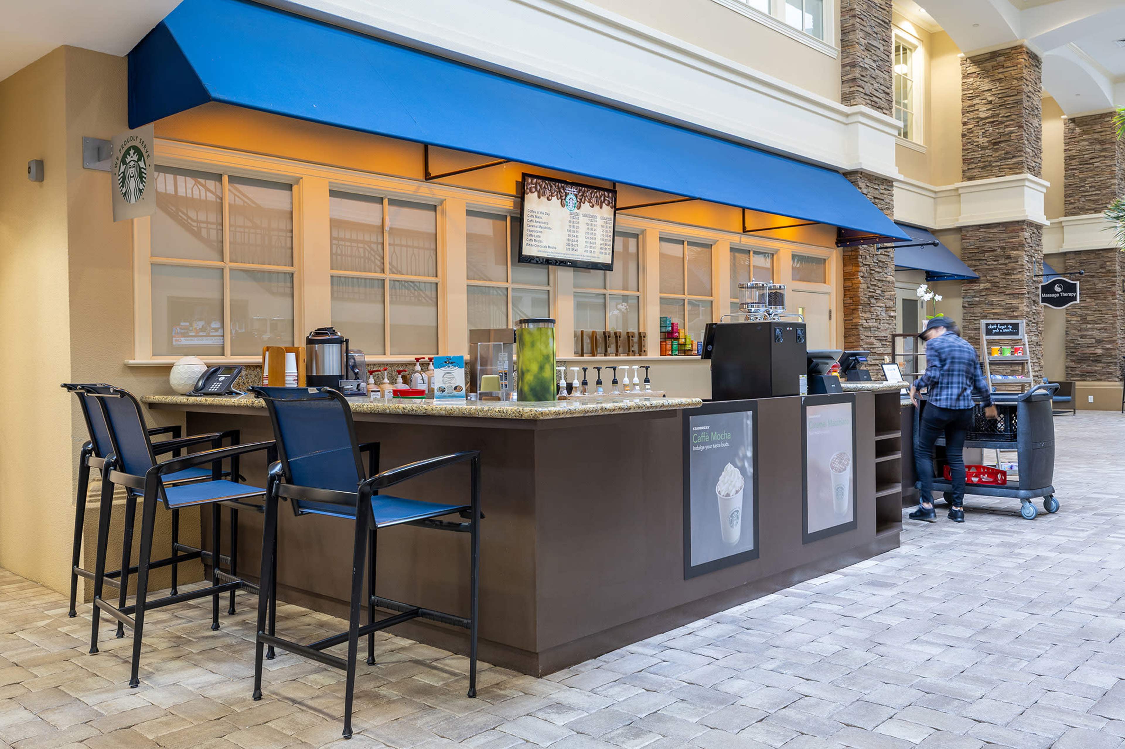 A coffee bar with a menu display and seating area is set up in a well-lit indoor space, alongside a person preparing a cart for service.