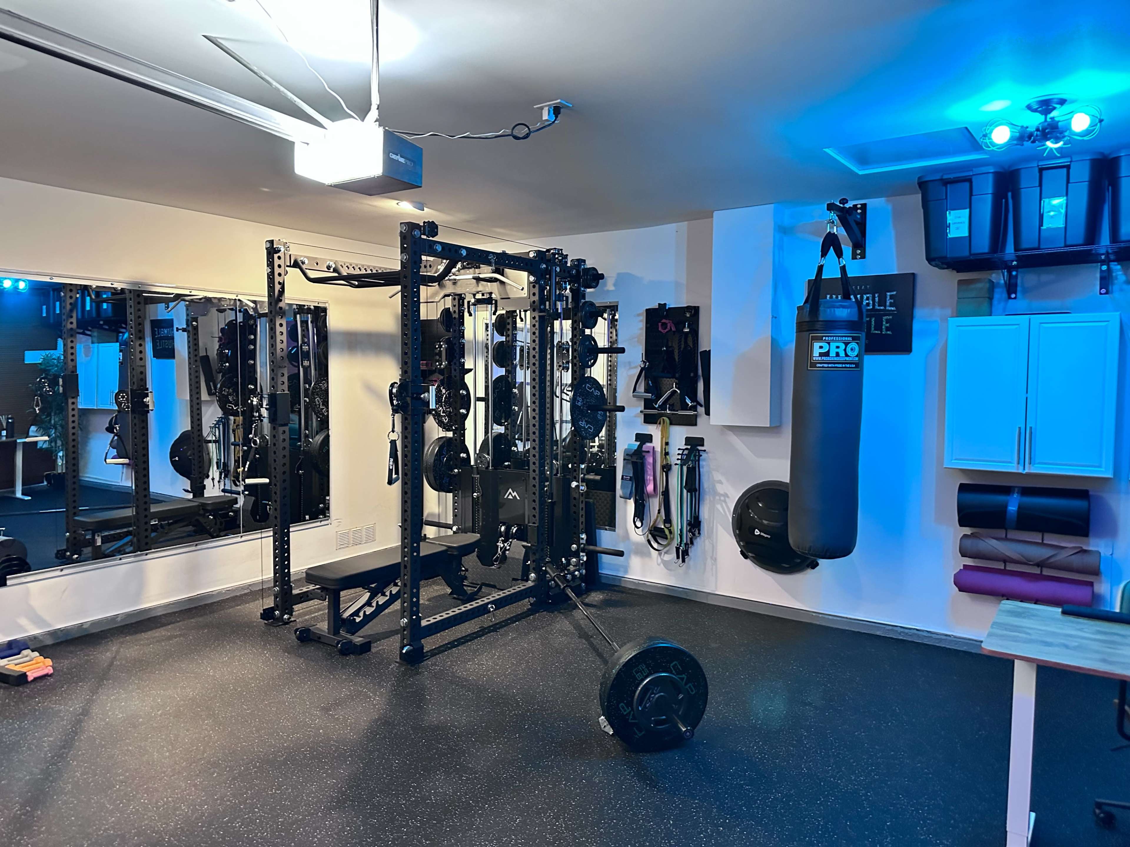 The image shows a well-equipped home gym featuring a power rack, mirrors, a heavy bag, and various fitness equipment against a black rubber floor.