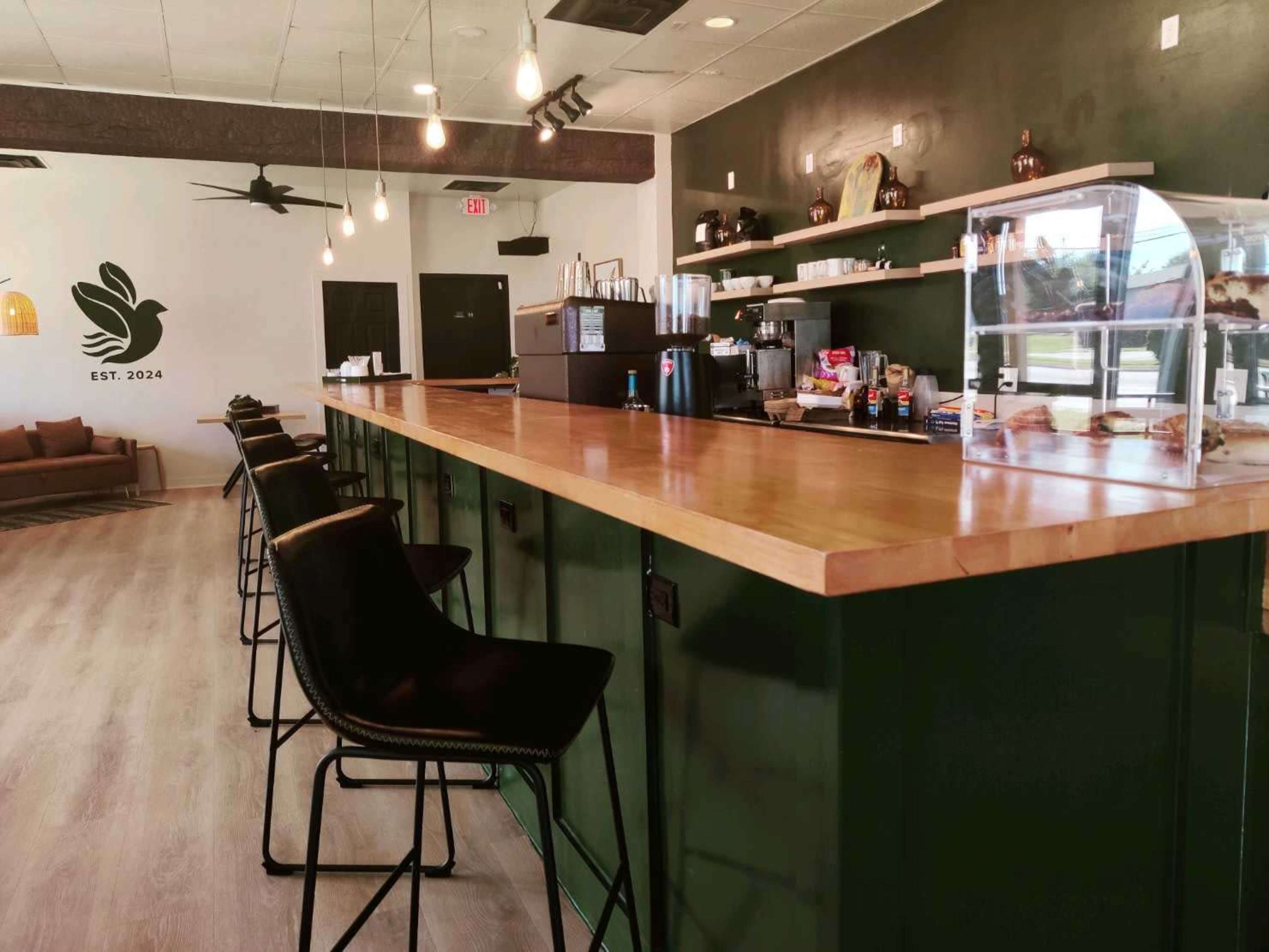 The image shows a modern café interior with a wooden bar counter, black high chairs, and a display case filled with pastries.