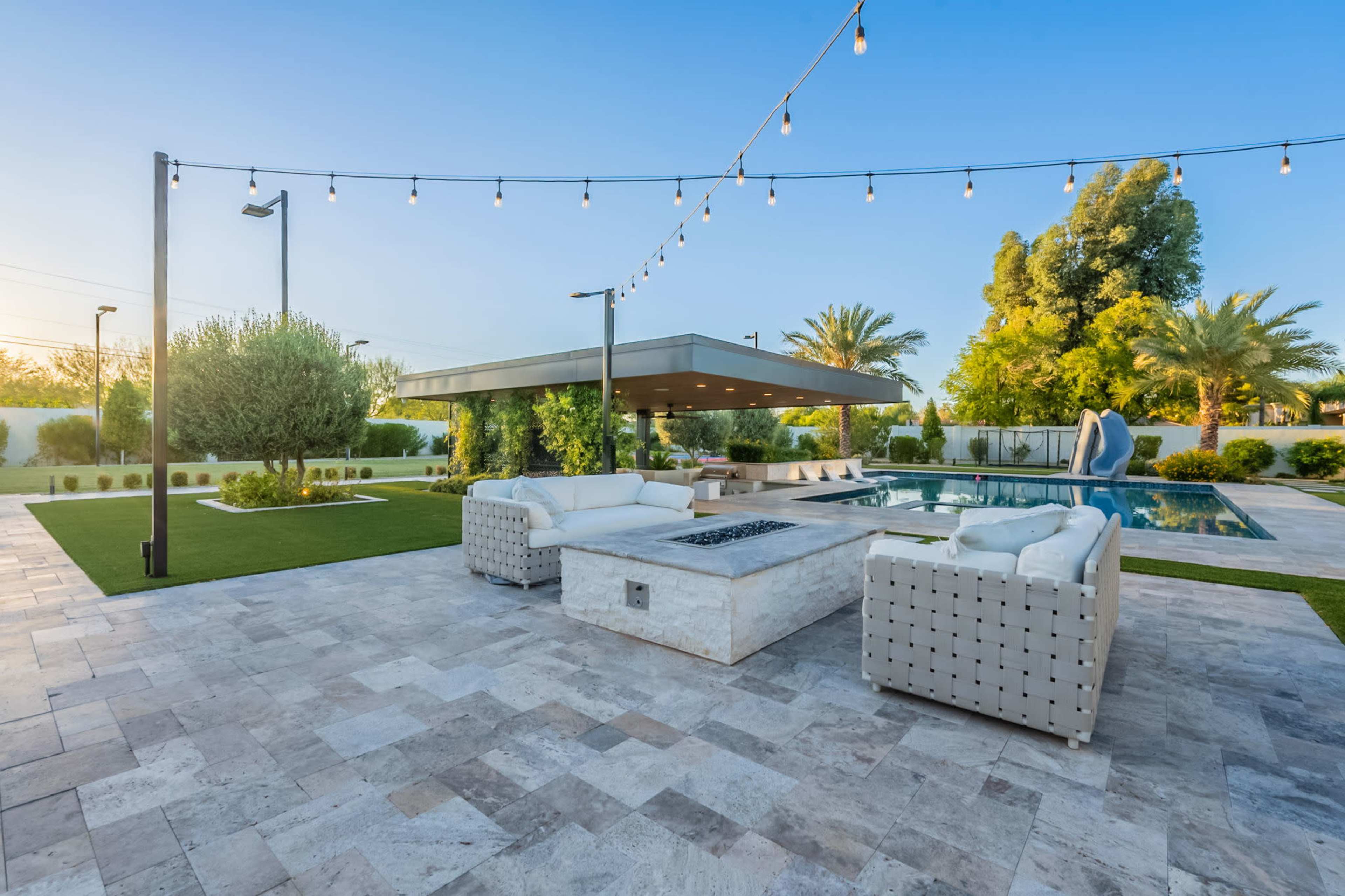 The image displays a modern backyard with a swimming pool, a seating area featuring a fire pit, and landscaping surrounded by palm trees.