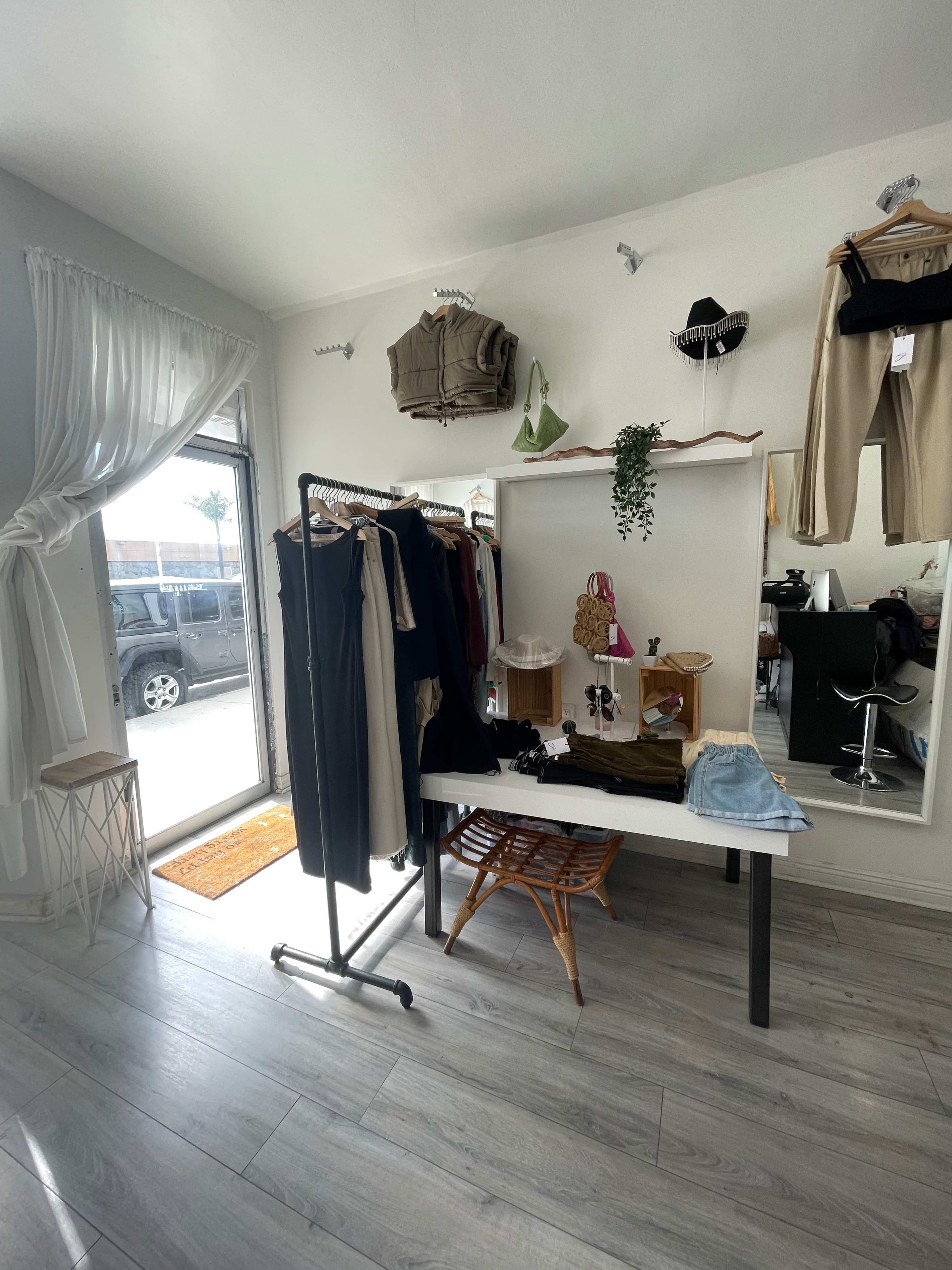 A clothing boutique with a rack of garments, a small display table, and a large mirror, illuminated by natural light from a nearby window.