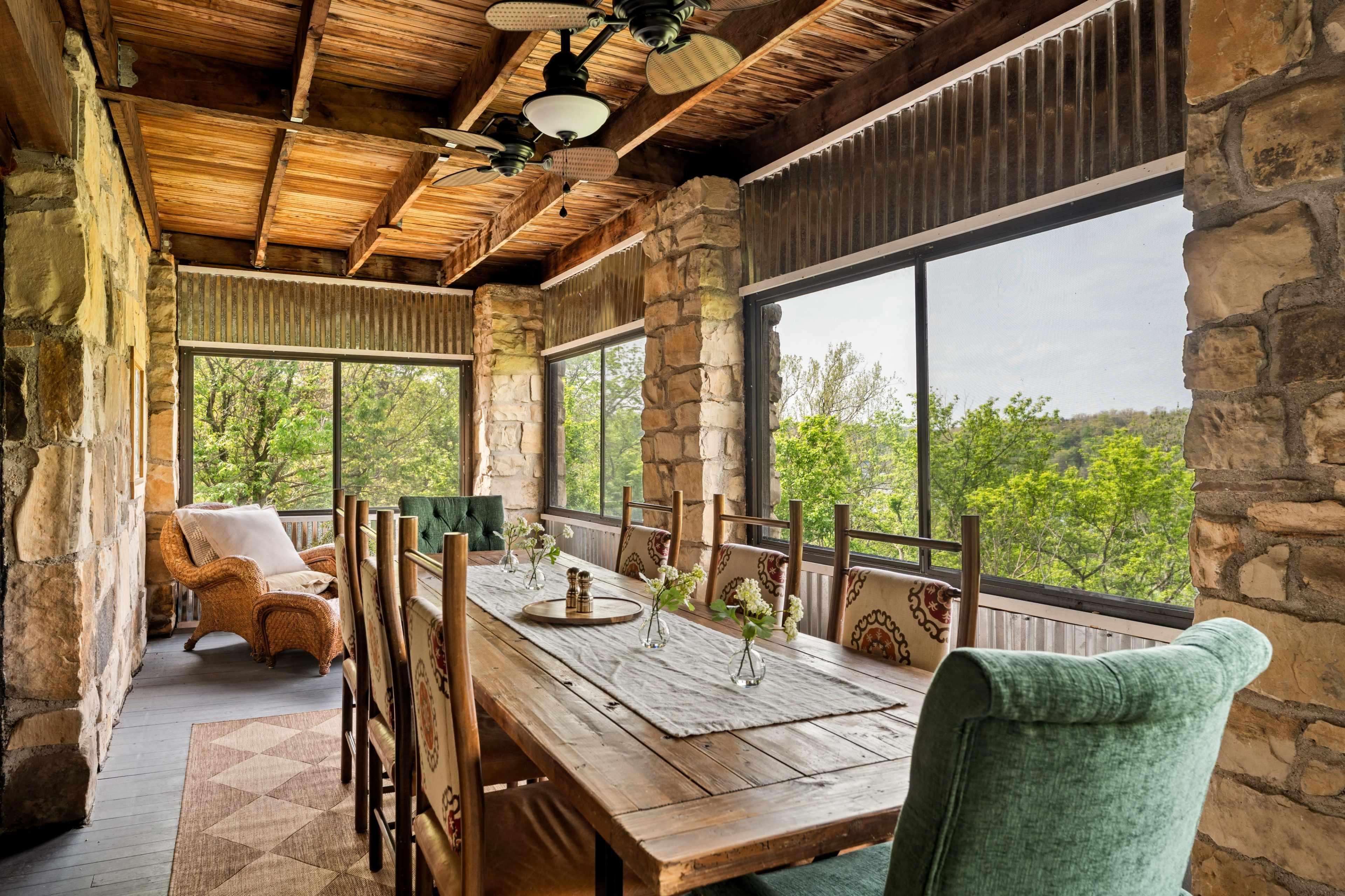 The image shows a rustic indoor porch area featuring a long wooden dining table surrounded by chairs, with stone walls and large windows revealing a view of trees outside.