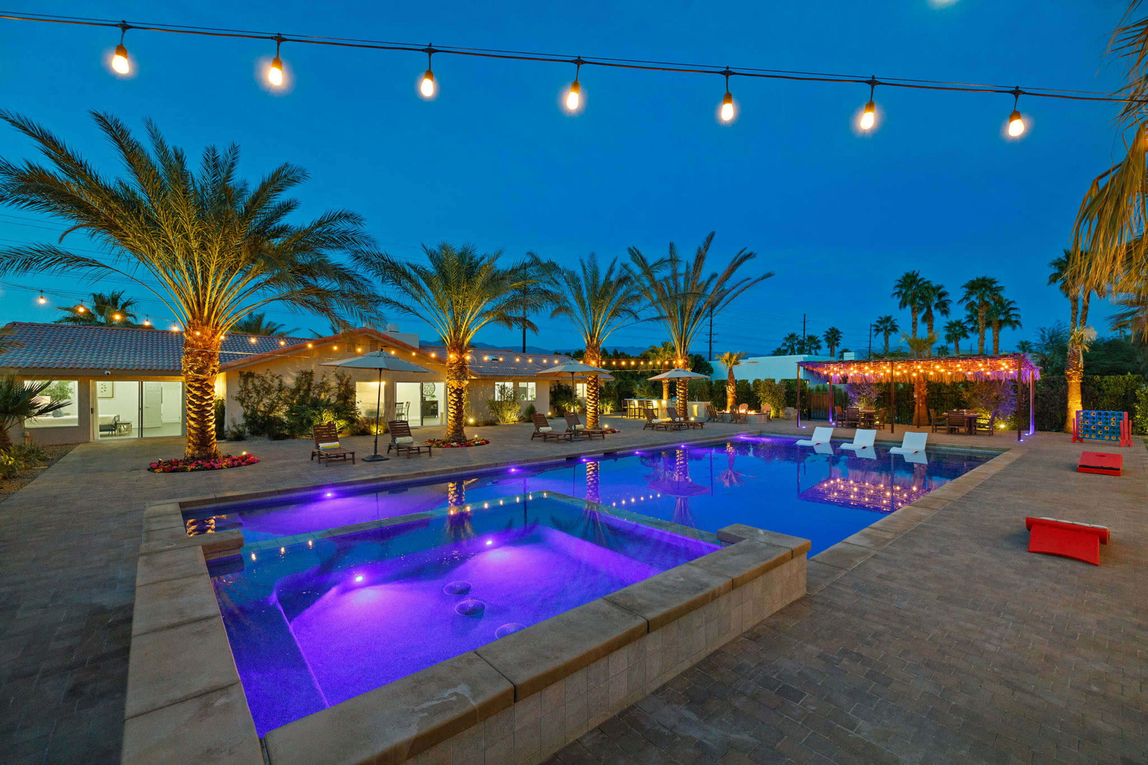 A backyard oasis features a swimming pool and spa, surrounded by palm trees and illuminated by string lights in the evening.