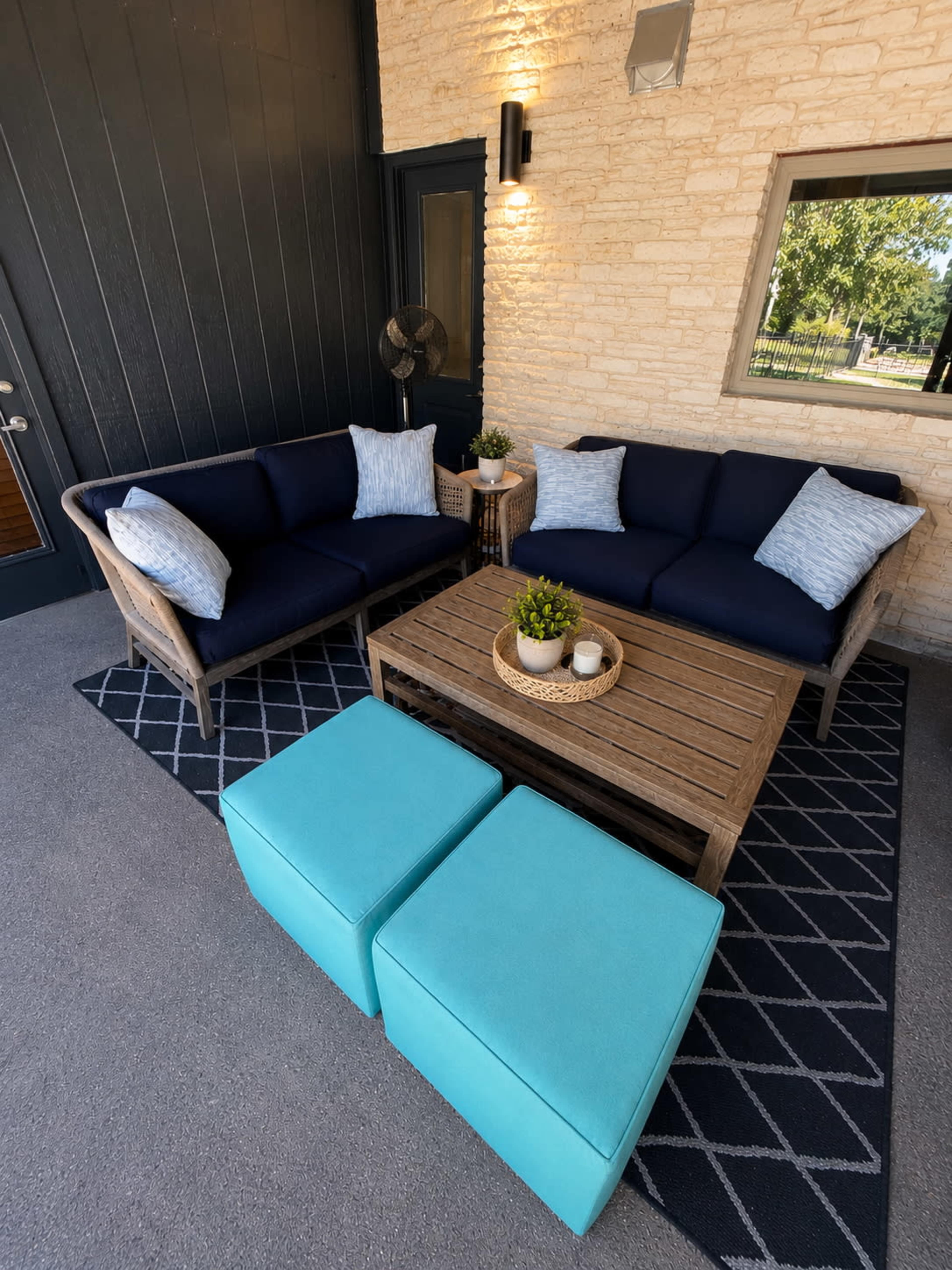 The image shows a cozy outdoor seating area with a dark blue couch, light blue accent pillows, a wooden coffee table, and two square turquoise ottomans on a geometric patterned rug.