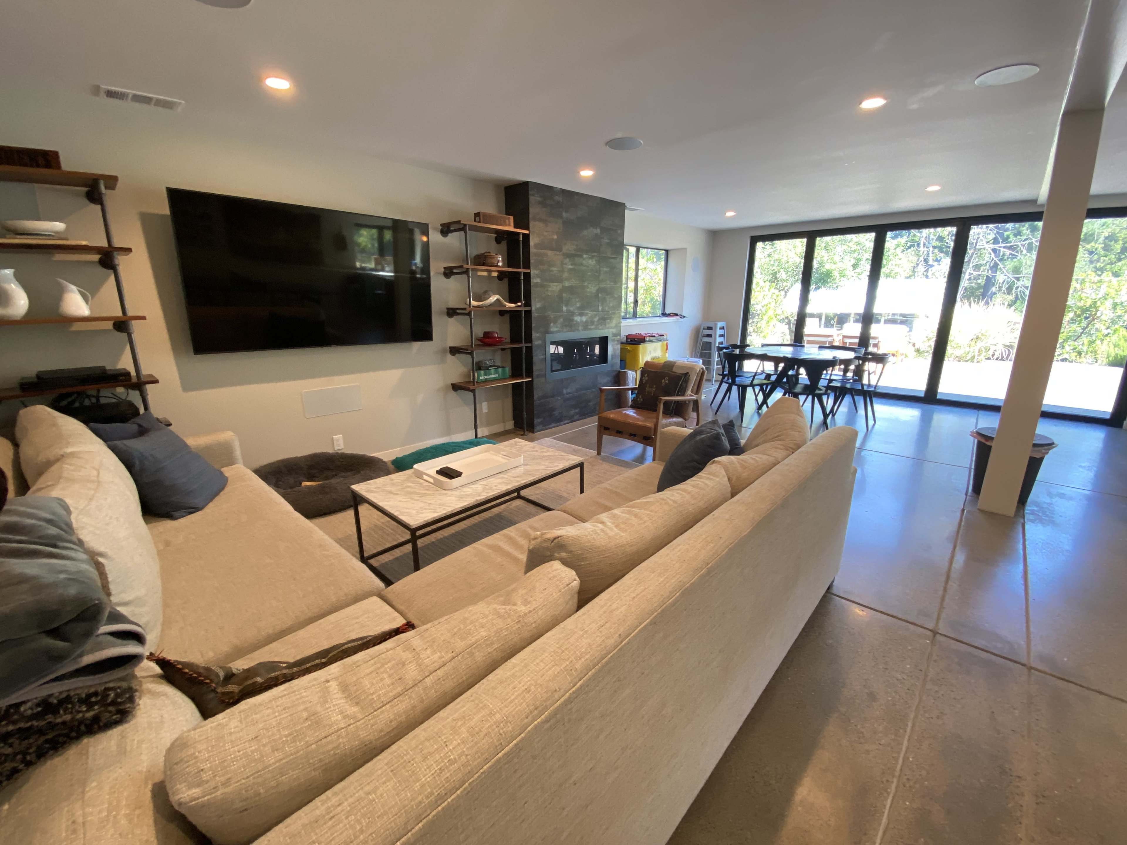 A modern living room with a large sectional sofa, a mounted television, shelves, and windows that provide a view of an outdoor area.