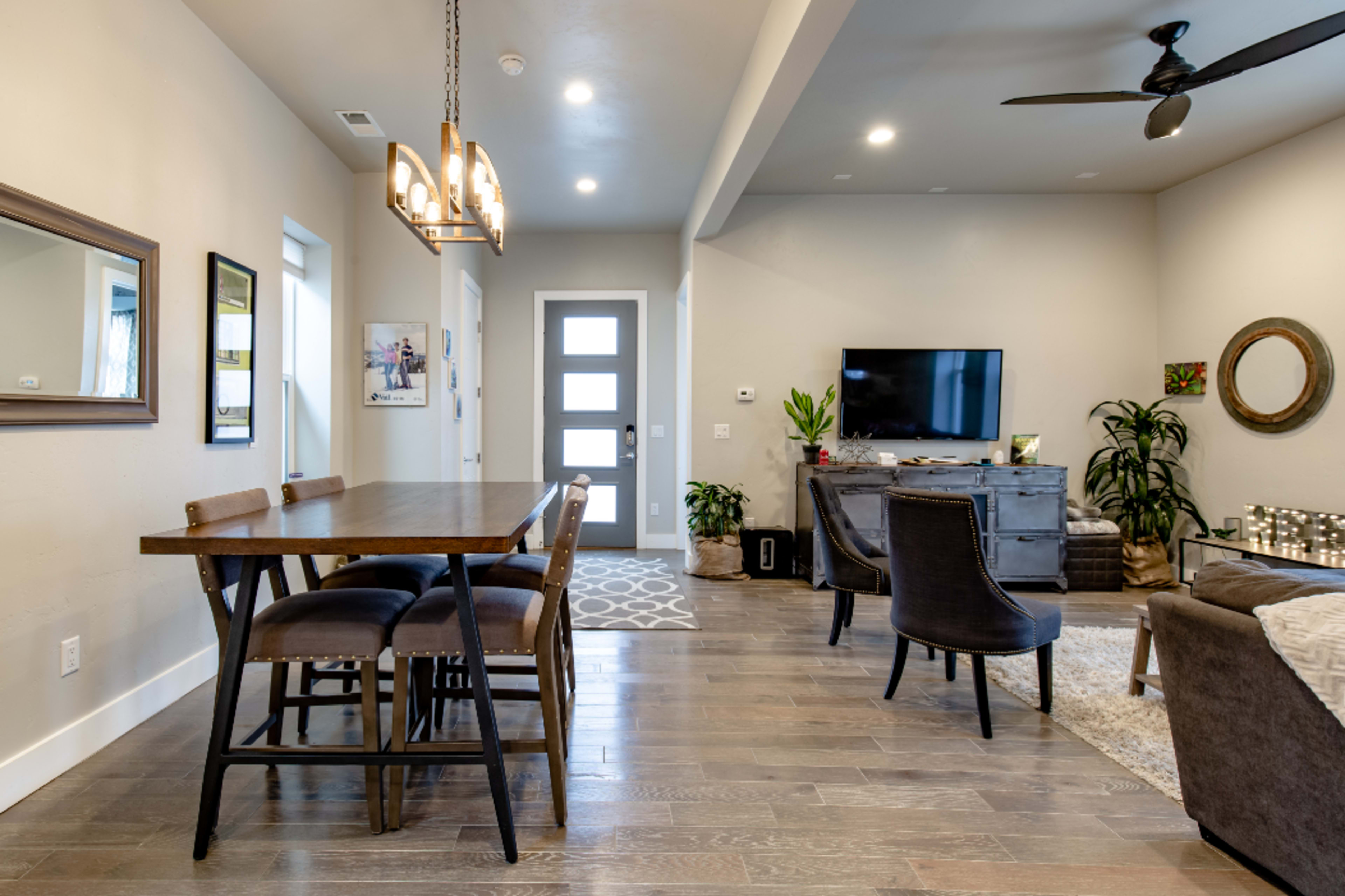 A modern living space with a dining area featuring a wooden table and chairs, a TV on a cabinet, and a mix of decorative plants and artwork on the walls.