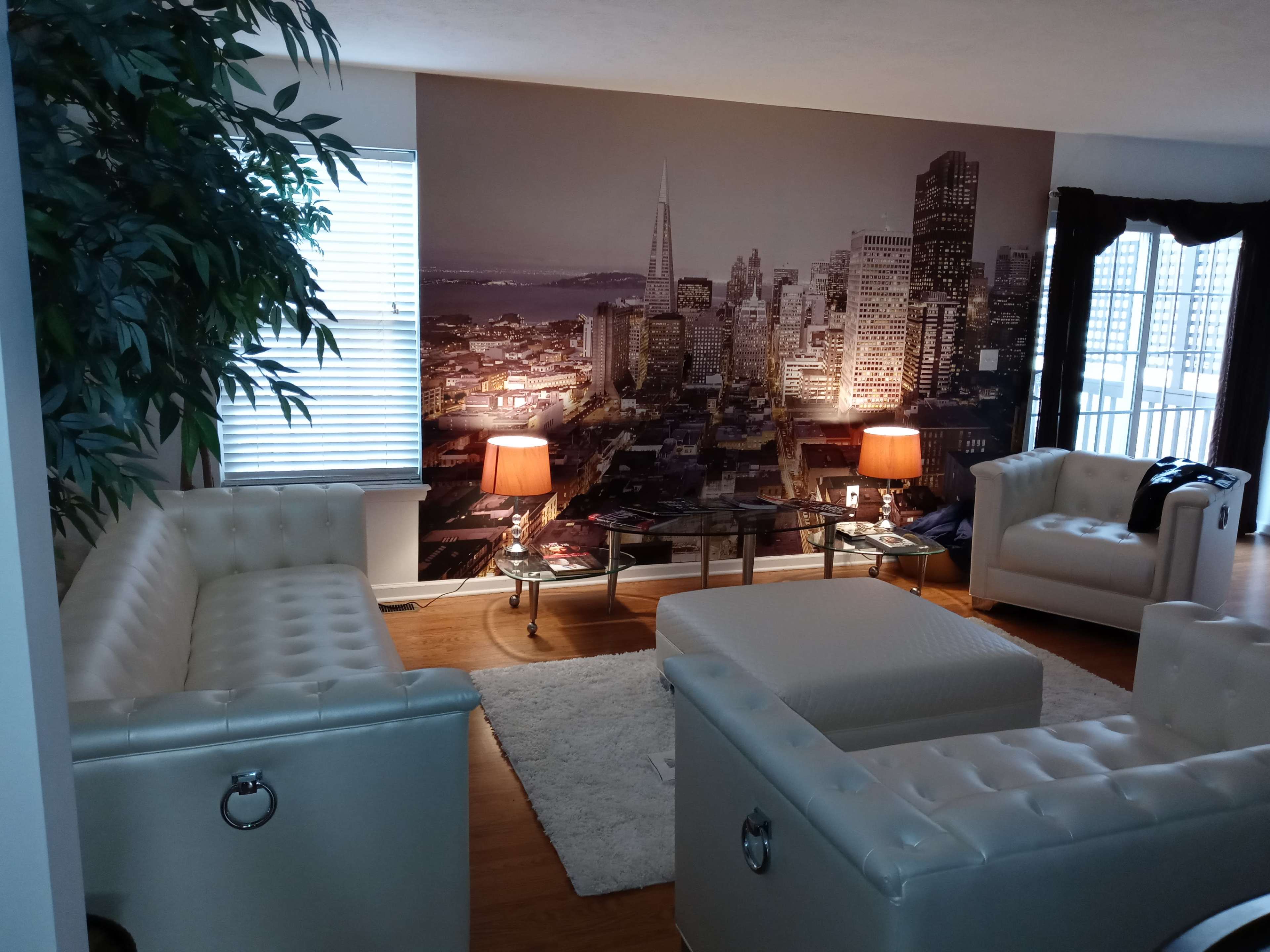 The image shows a modern living room with white leather furniture, a glass coffee table, and a large wall mural depicting a city skyline at night.