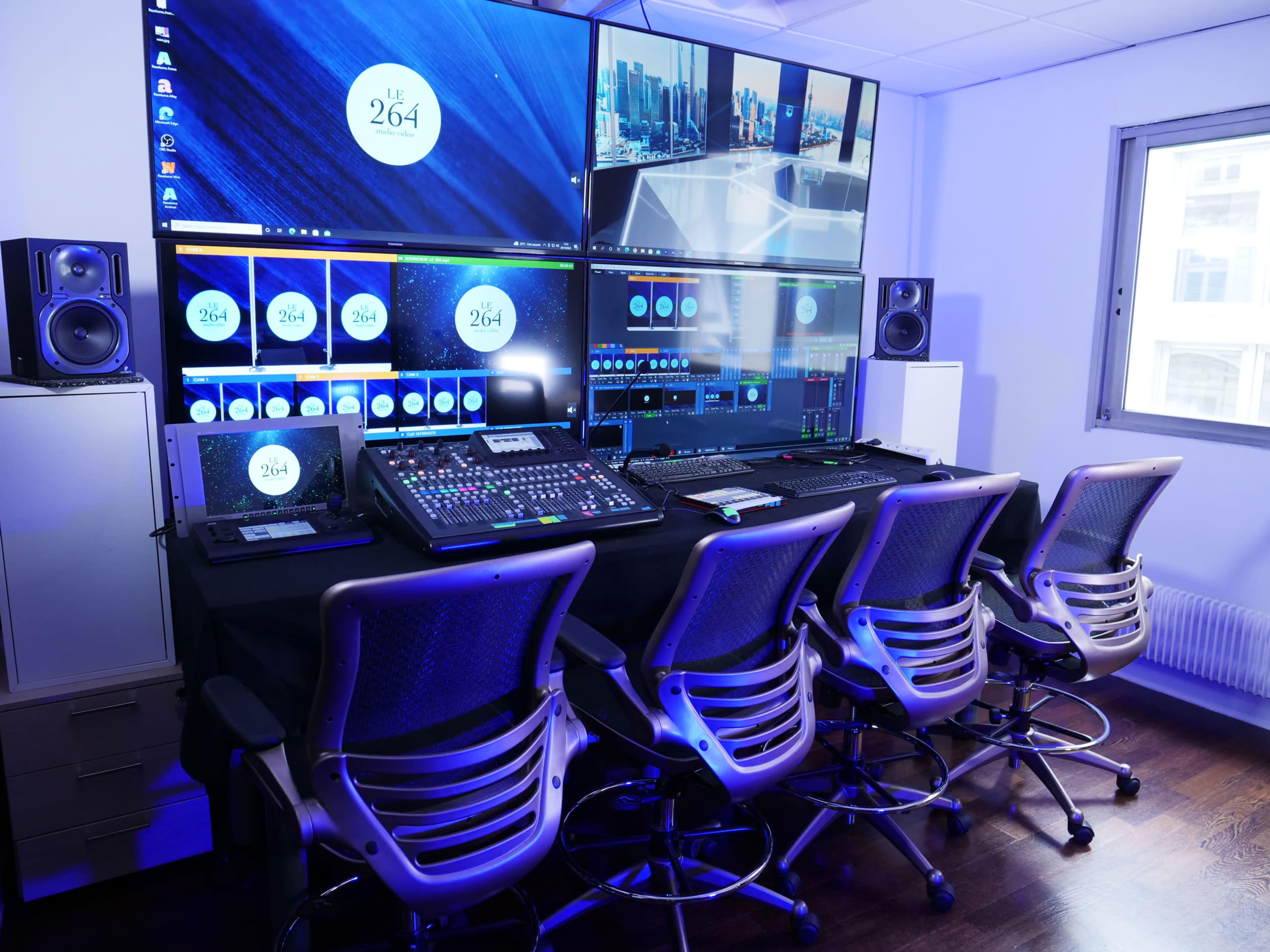 The image shows a modern control room with multiple large screens, audio equipment, and rows of swivel chairs arranged in front of a desk.