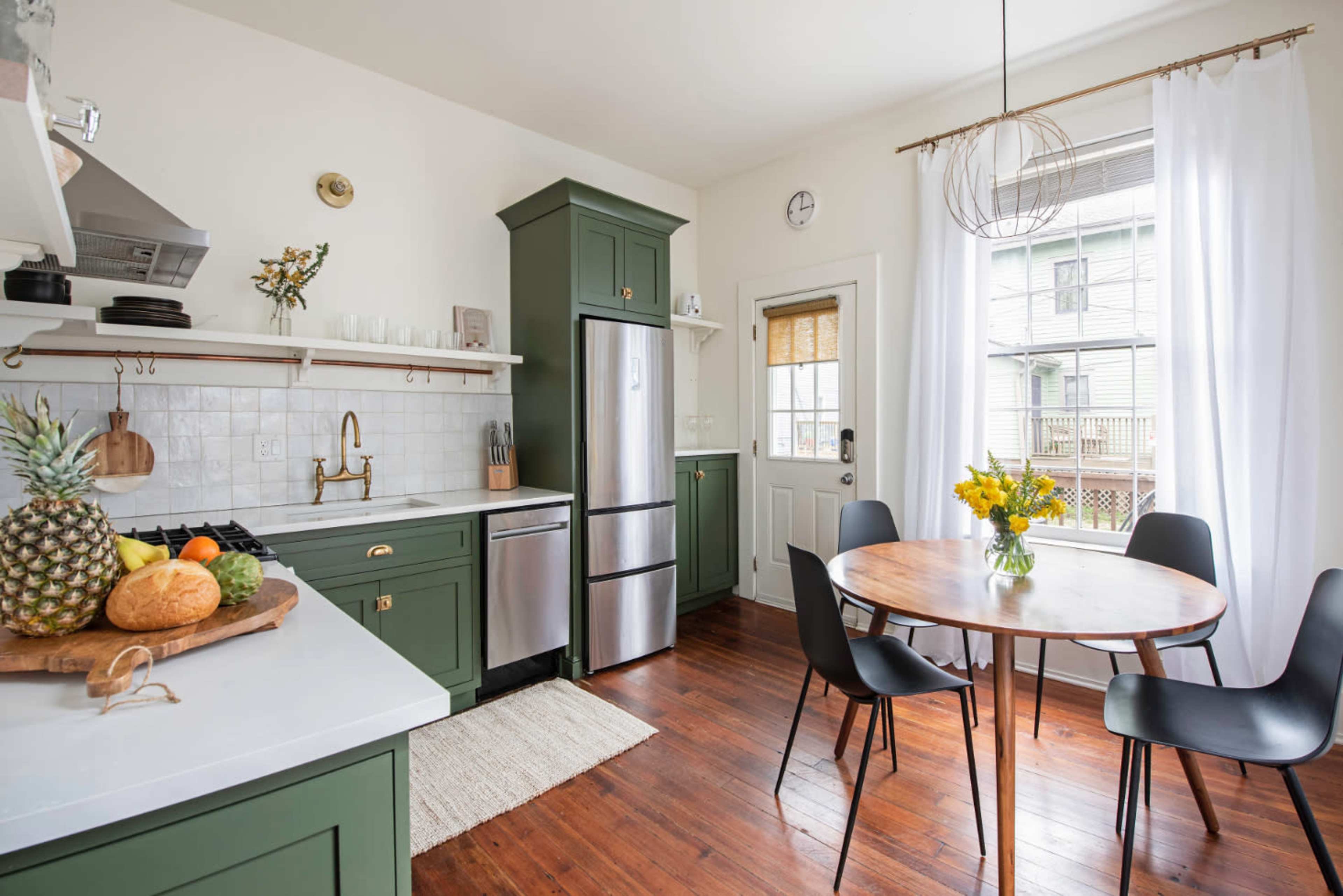 The image shows a compact kitchen featuring green cabinetry, a stainless steel refrigerator, and a wooden dining table with black chairs.