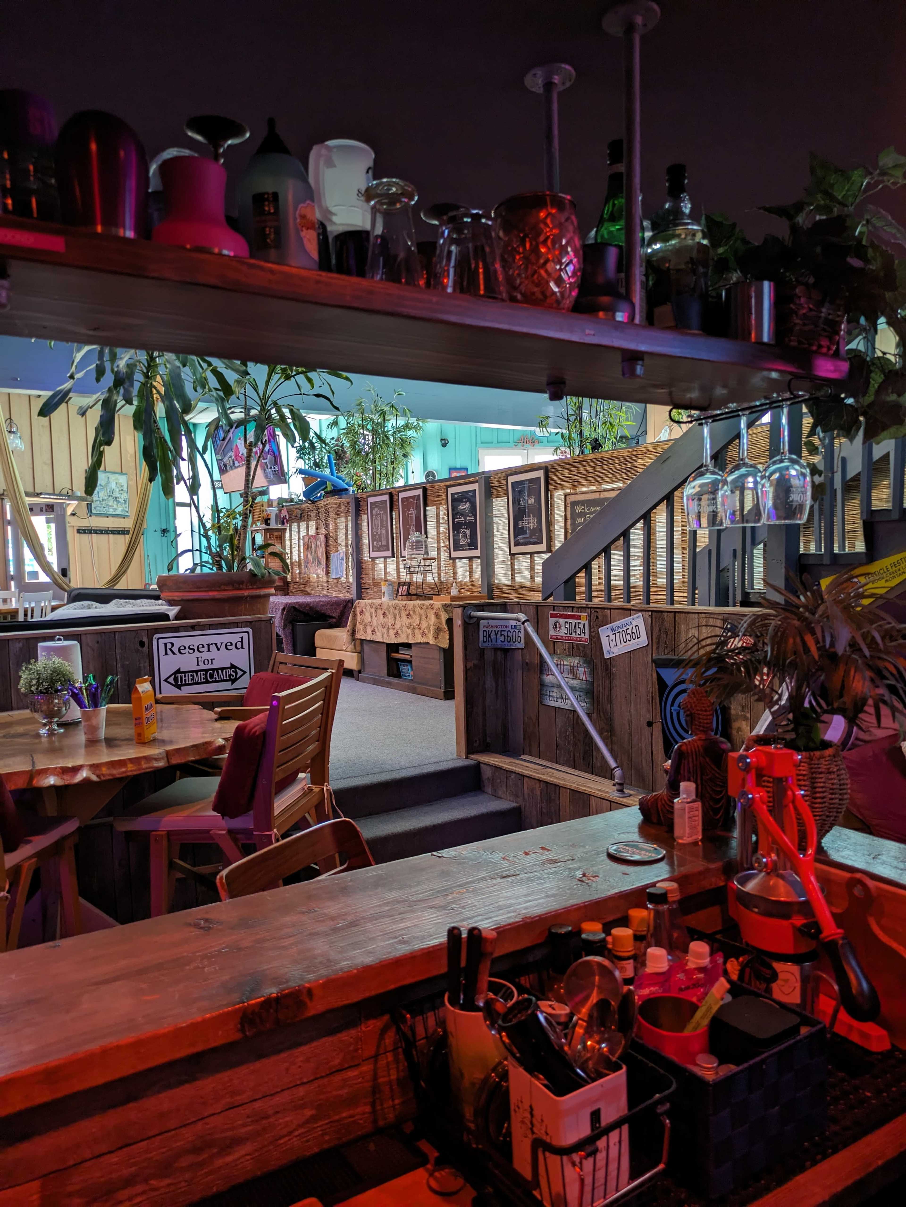 A bar area with various glassware and utensils on display, overlooking a reserved seating section decorated with plants and framed photographs.