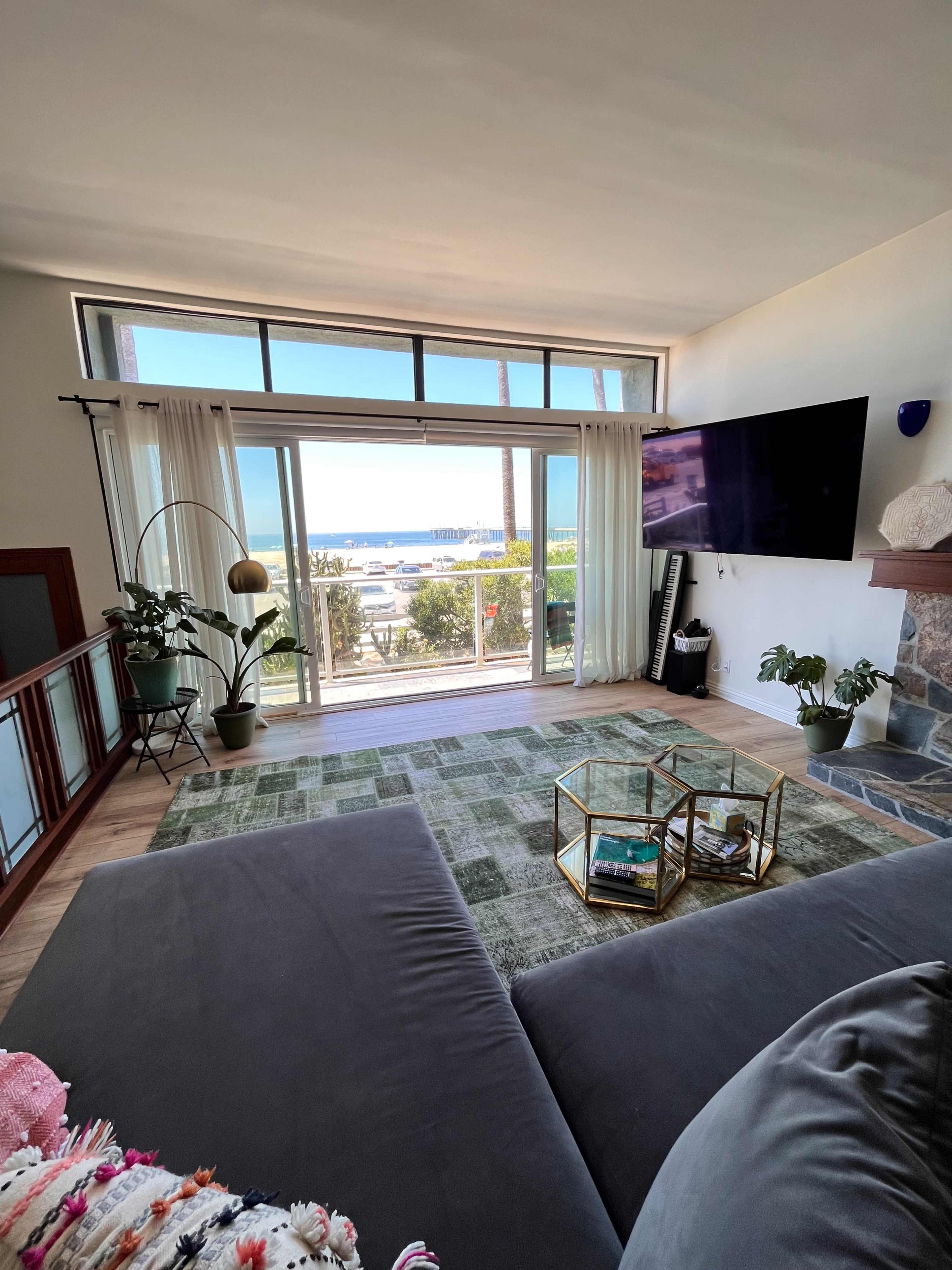 The living room features large windows with a view of the ocean, a gray sofa, a green rug, and a stone fireplace.