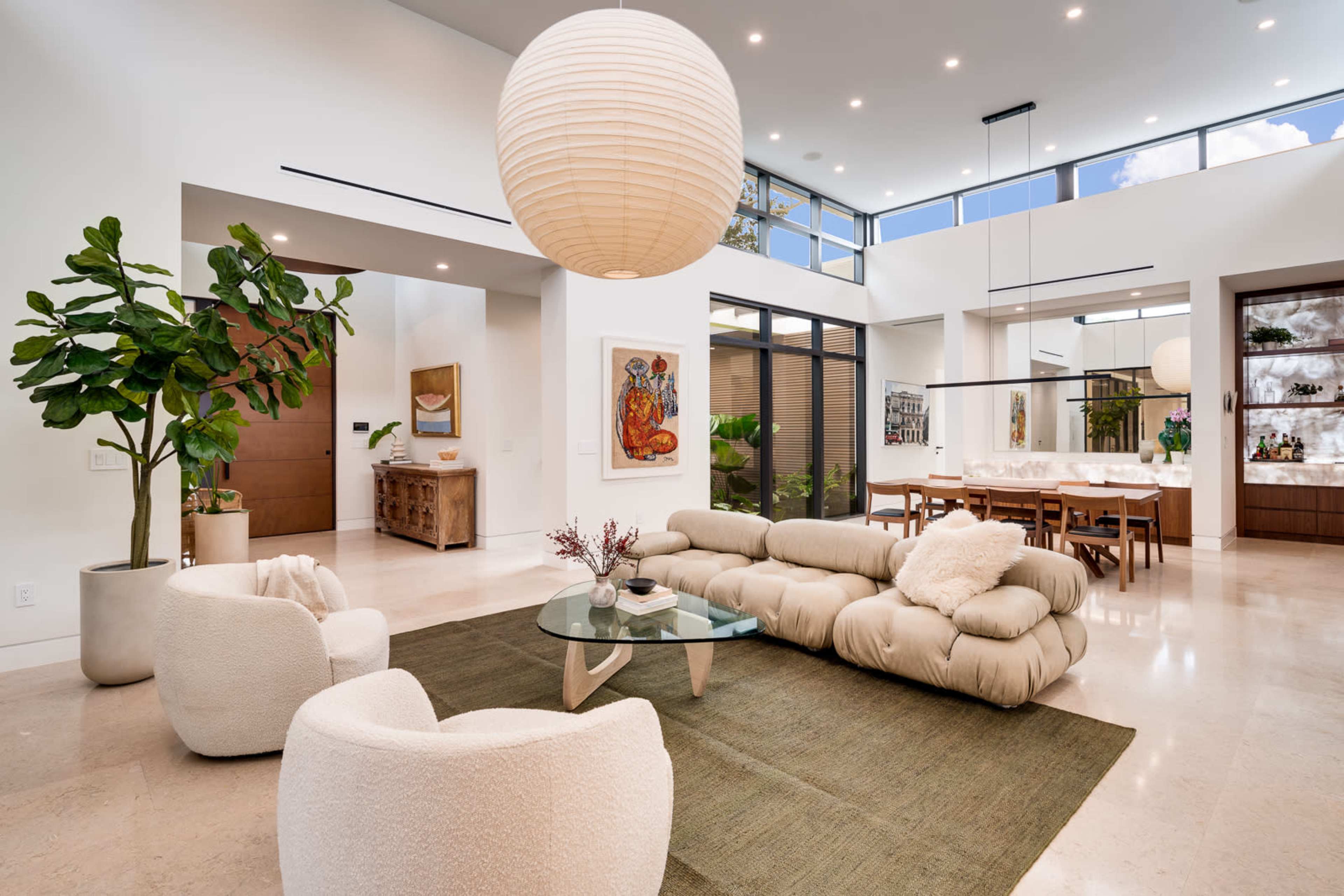 A modern open-concept living area featuring a large rug, a glass coffee table, a plush sofa, two accent chairs, a dining area with a wooden table, and large windows allowing natural light.