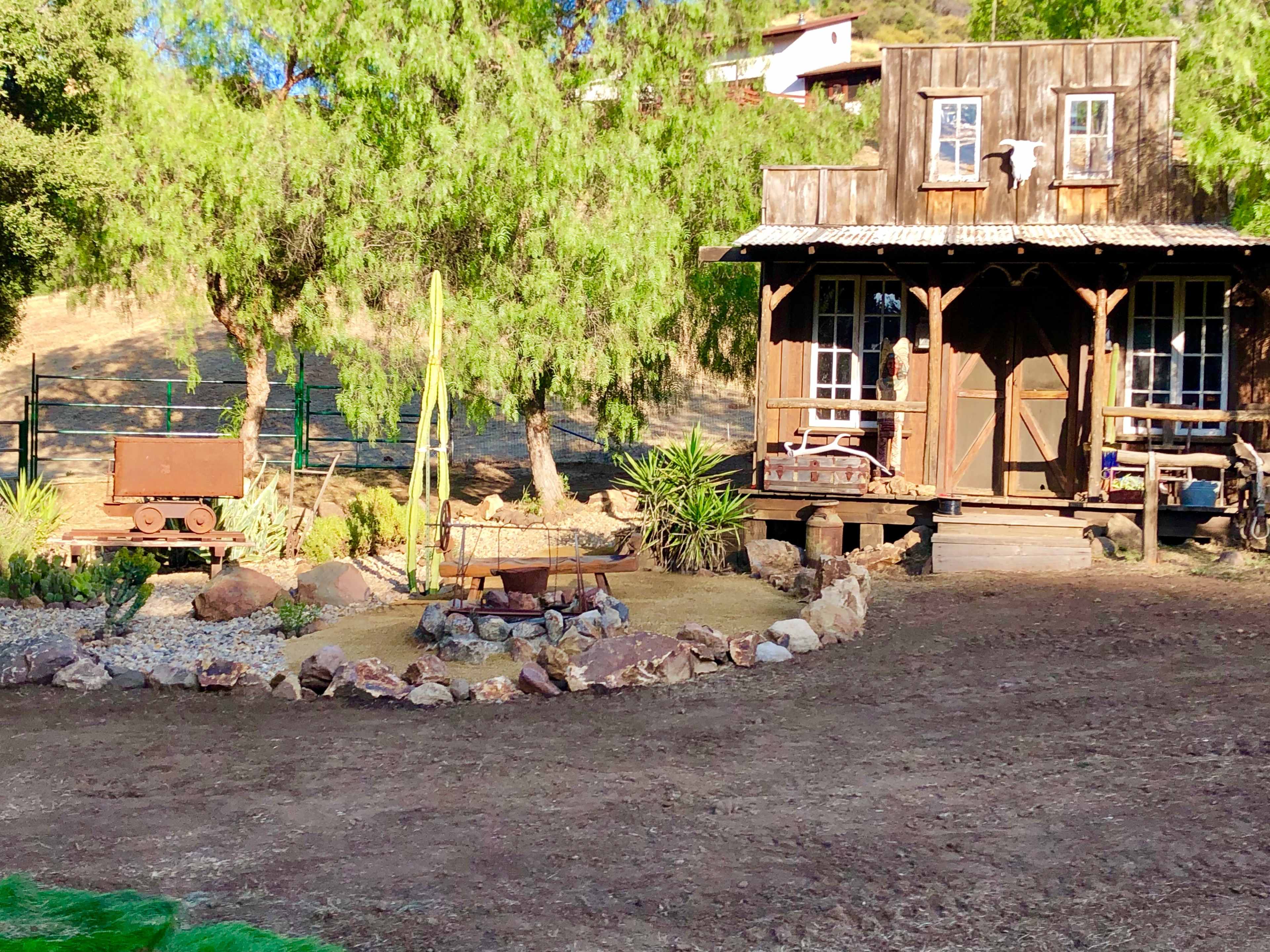 A rustic wooden cabin surrounded by landscaped gravel and stone pathways, with a small wagon and circular fire pit in the foreground.