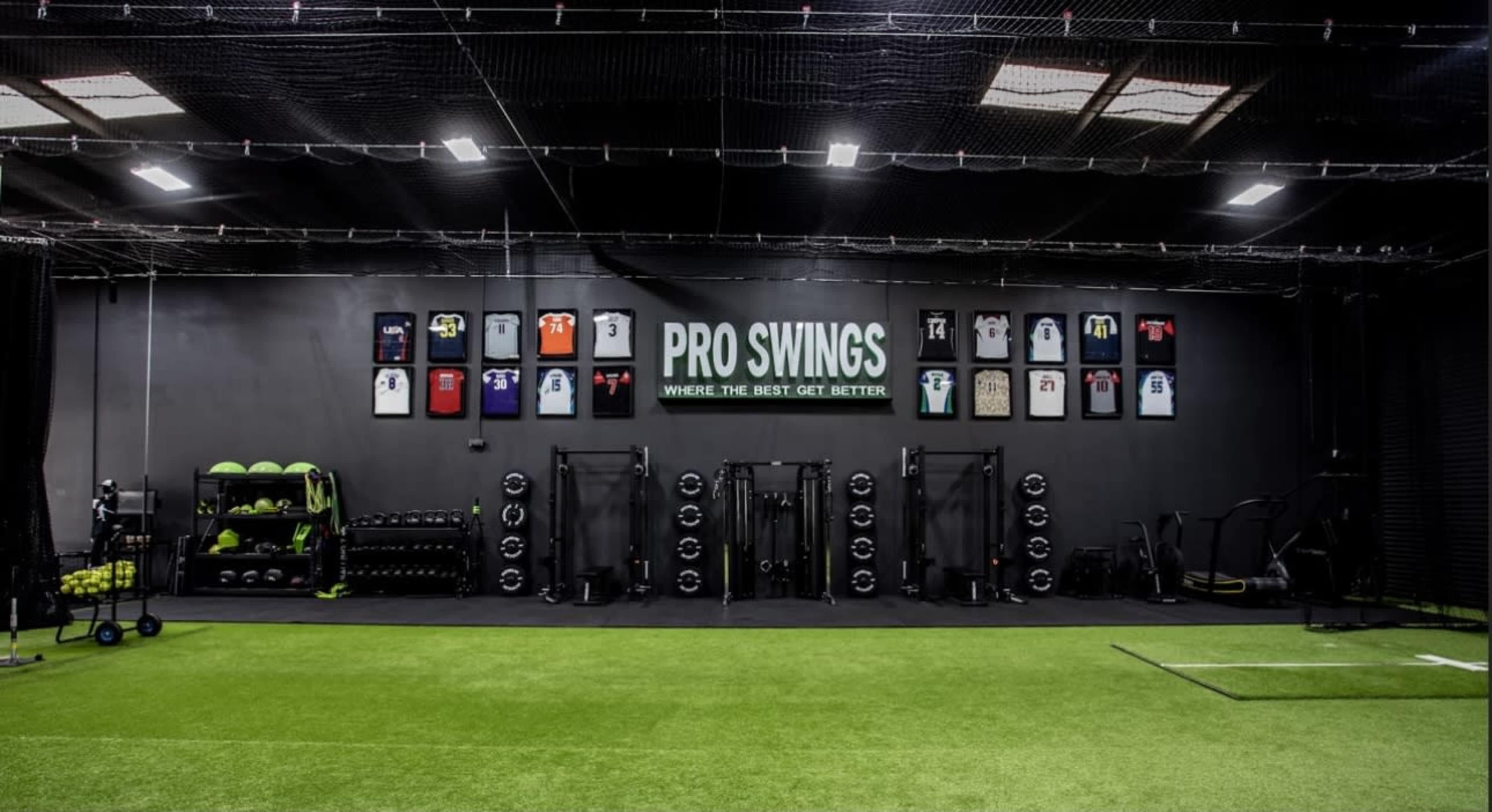 The image shows a modern gym space with turf flooring, wall-mounted jerseys hung on a dark wall, and various strength training equipment arranged on one side.