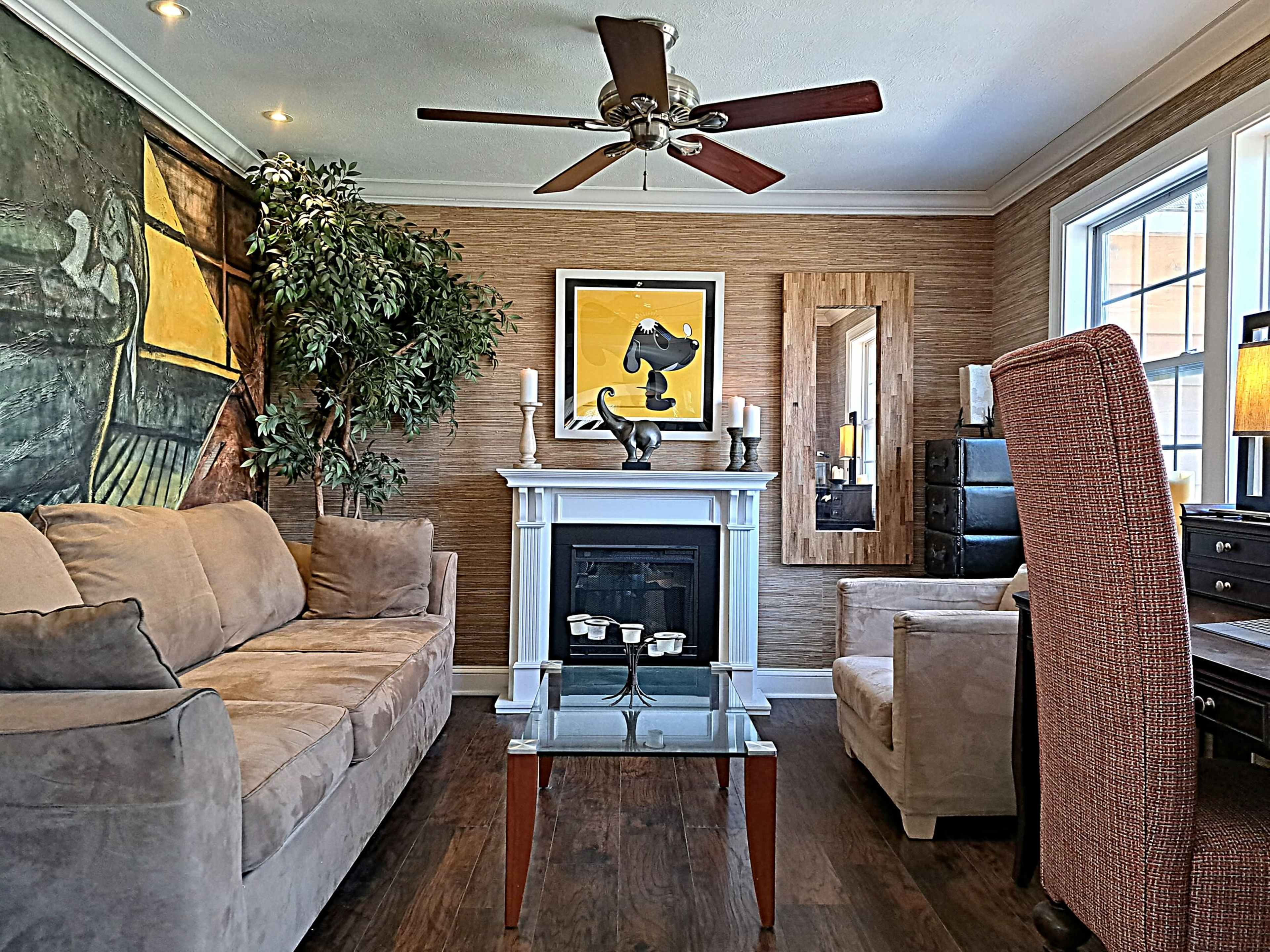 A living room features a beige sofa, a glass coffee table, a fireplace, and artwork of a cat, surrounded by indoor plants and a wooden mirror.
