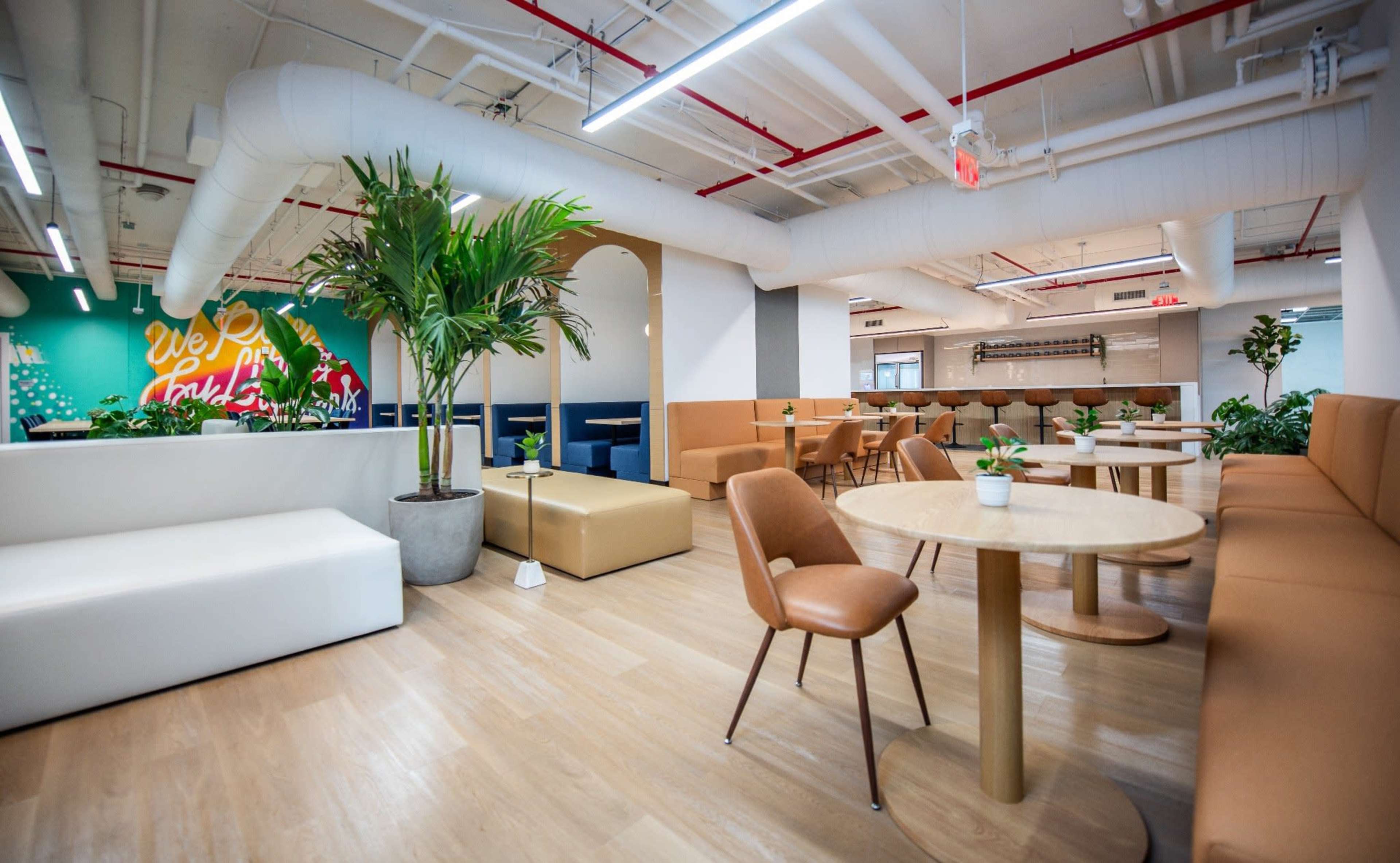 The image shows a modern office lounge area with seating, tables, and decorative plants, featuring a colorful mural on the wall.