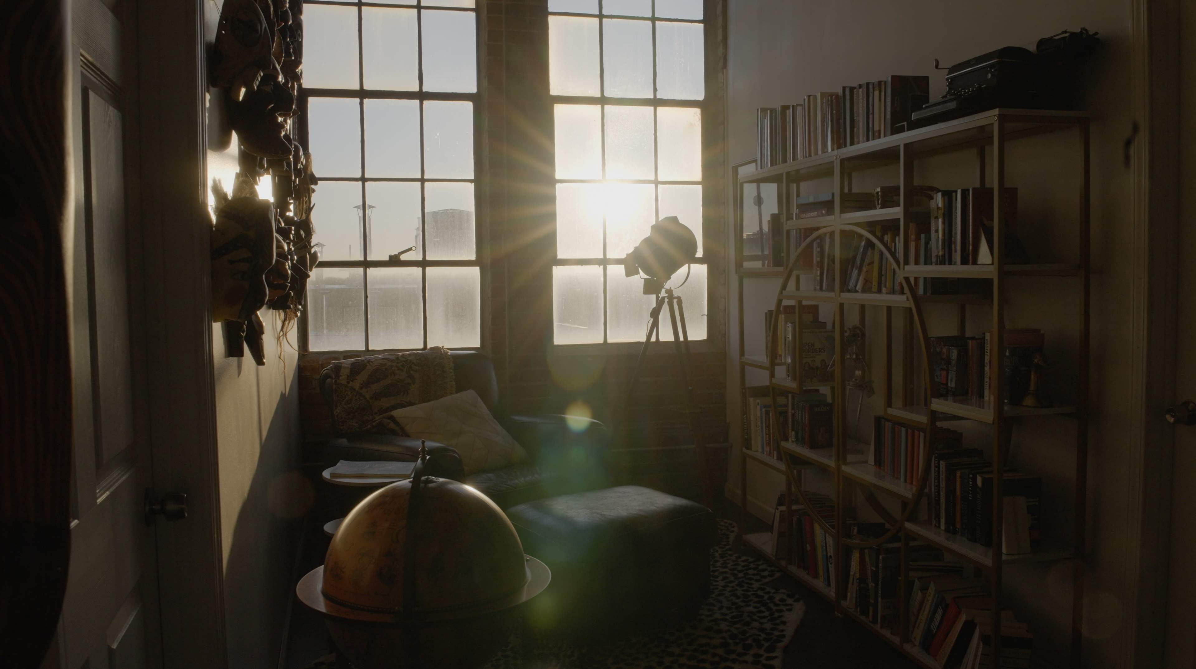 A sunlit room features large windows, a globe, a bookshelf filled with books, and a cozy seating area next to a modern tripod lamp.
