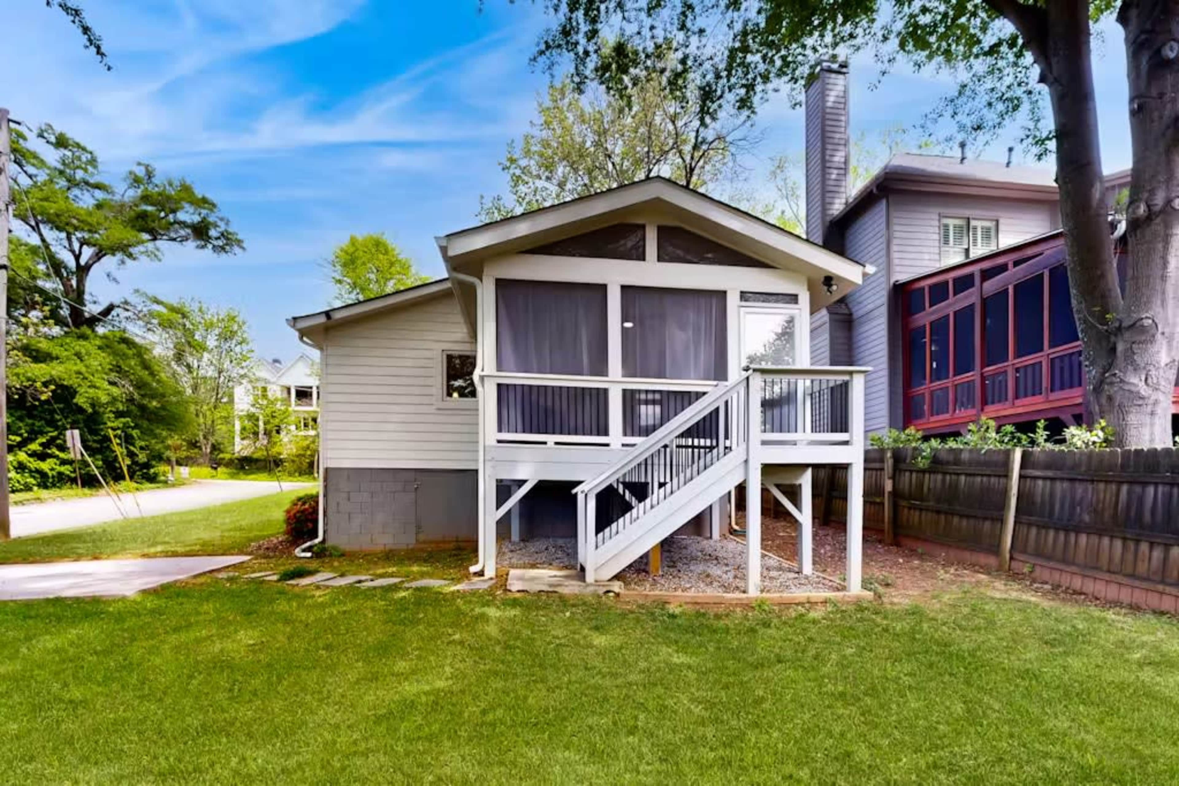 The image shows a small gray house with a porch and white railings, situated on a grassy lawn next to a tree-lined road.