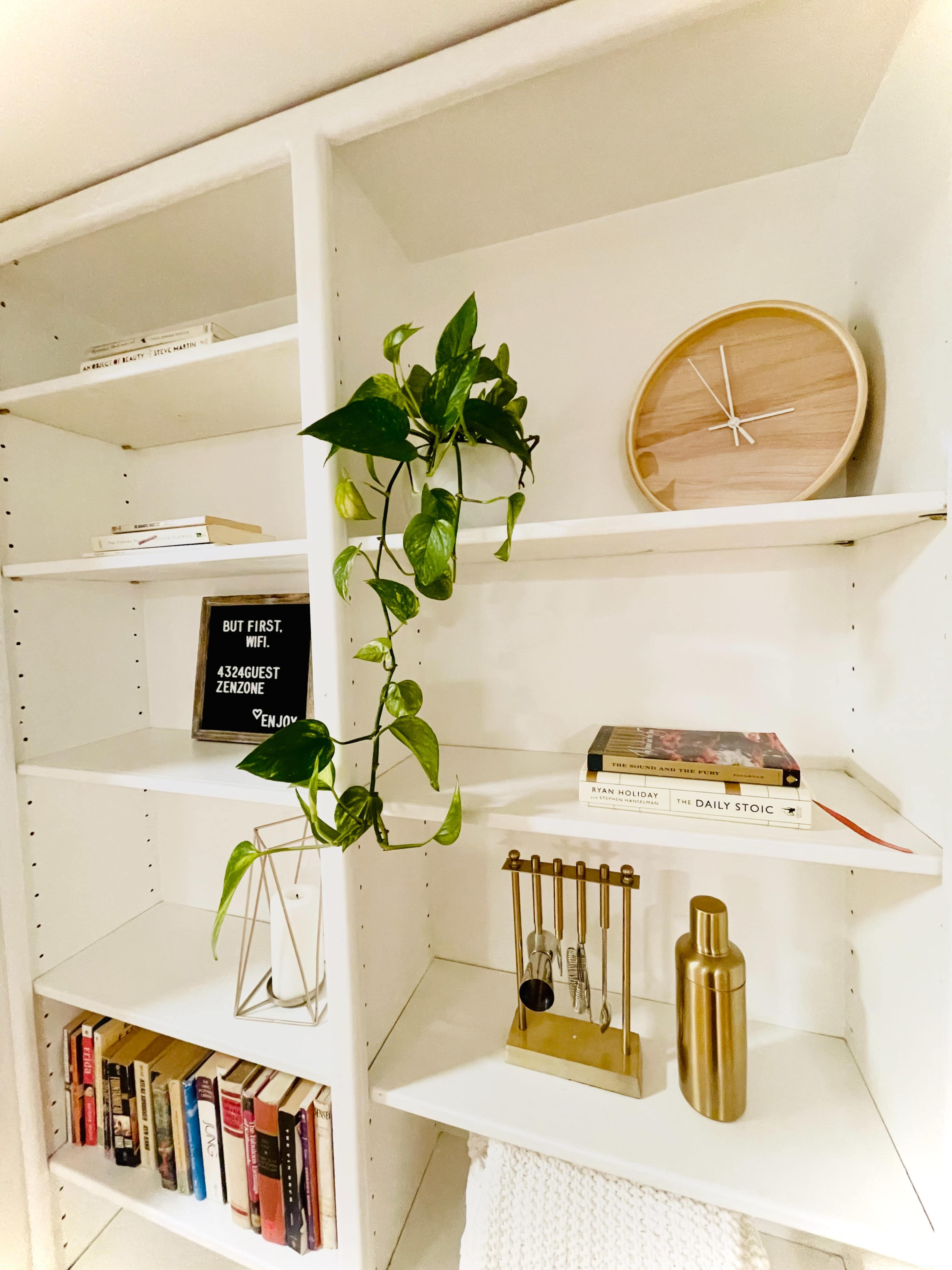 A neatly organized bookshelf features books, decorative items, a clock, a plant, and a small metal sculpture.