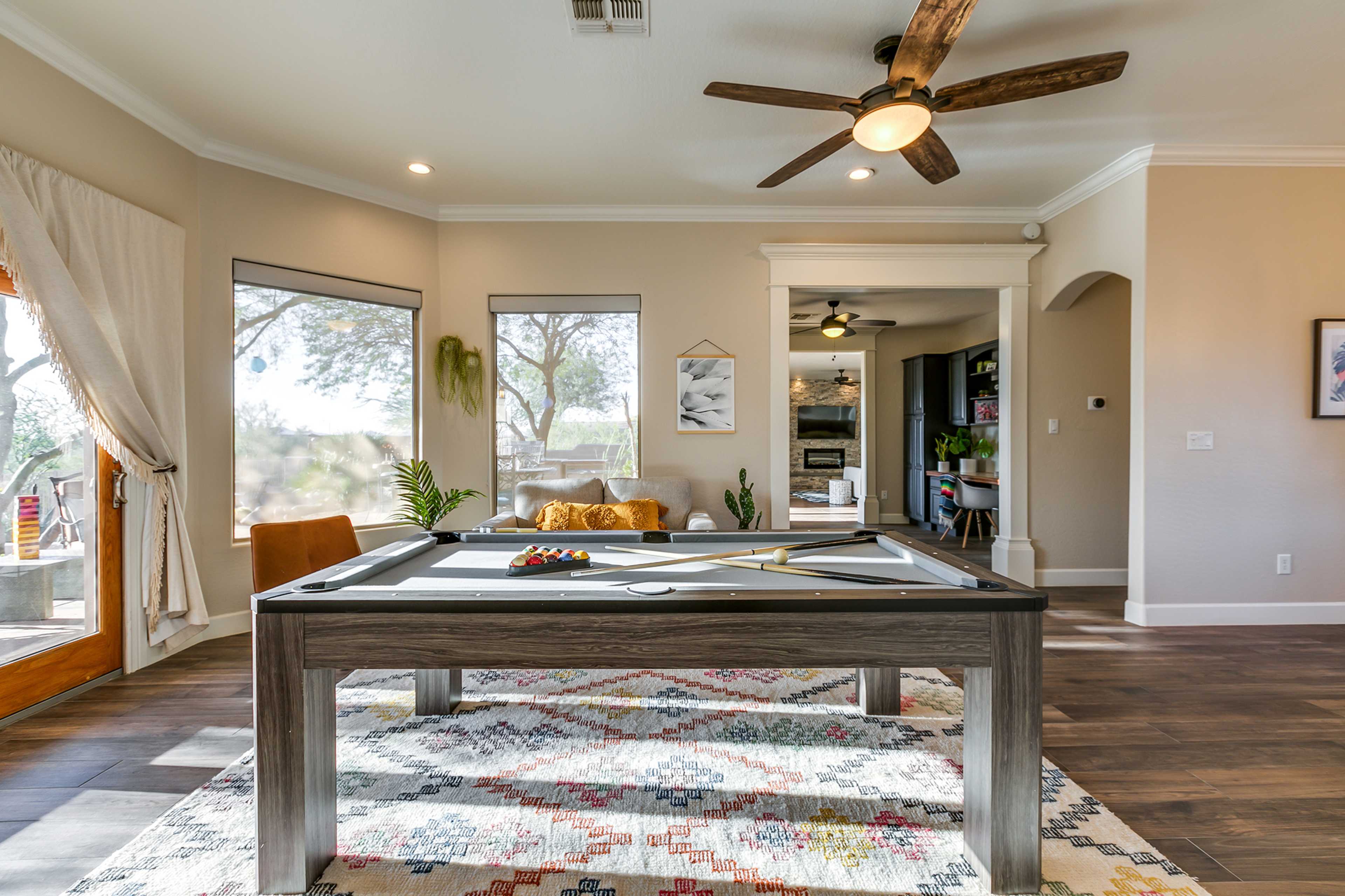 A well-lit room featuring a pool table in the center, surrounded by large windows and a seating area.