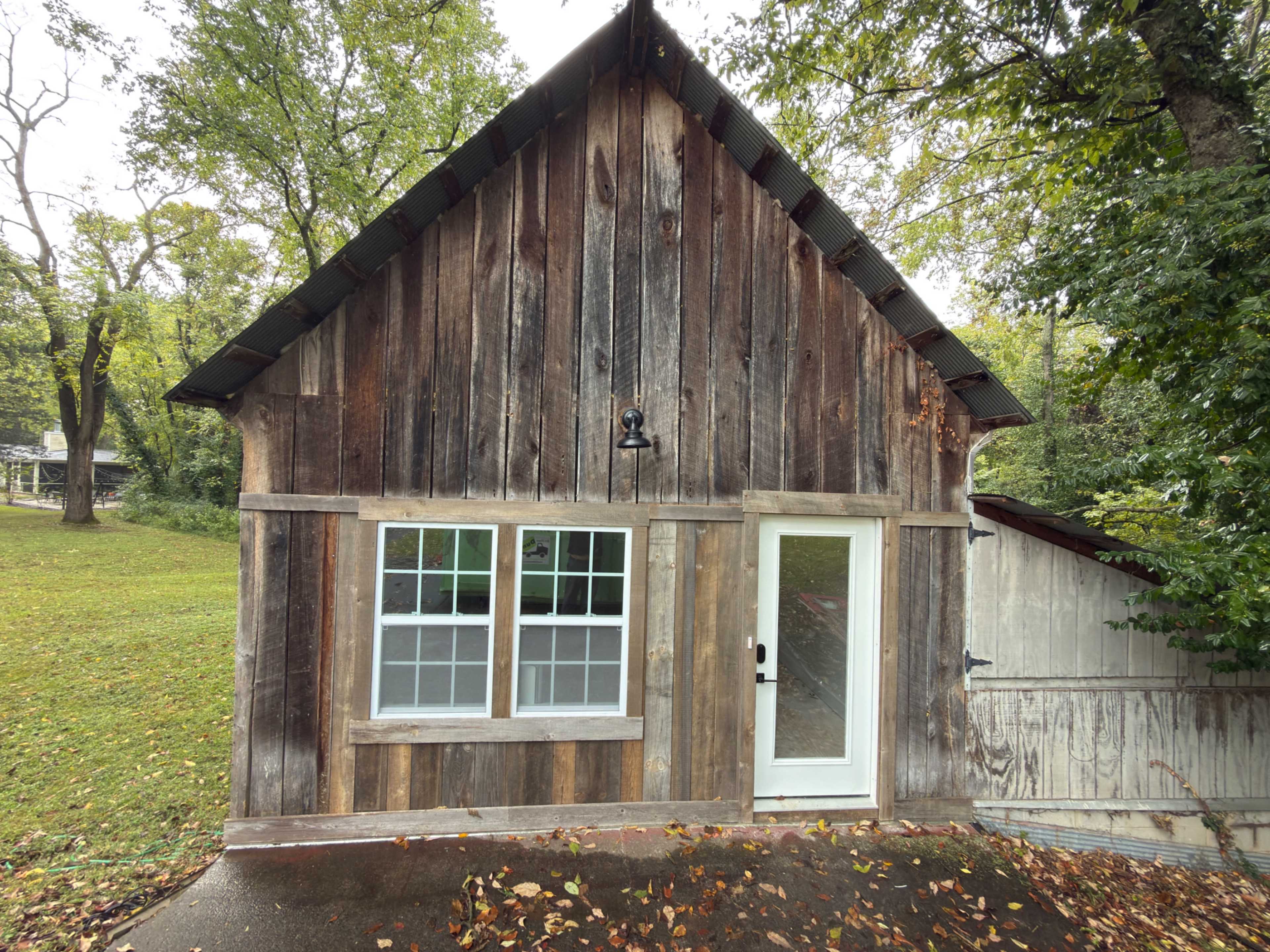 The image shows a rustic wooden cabin with a sloped roof, featuring two windows and a white door, set against a backdrop of trees and grass.