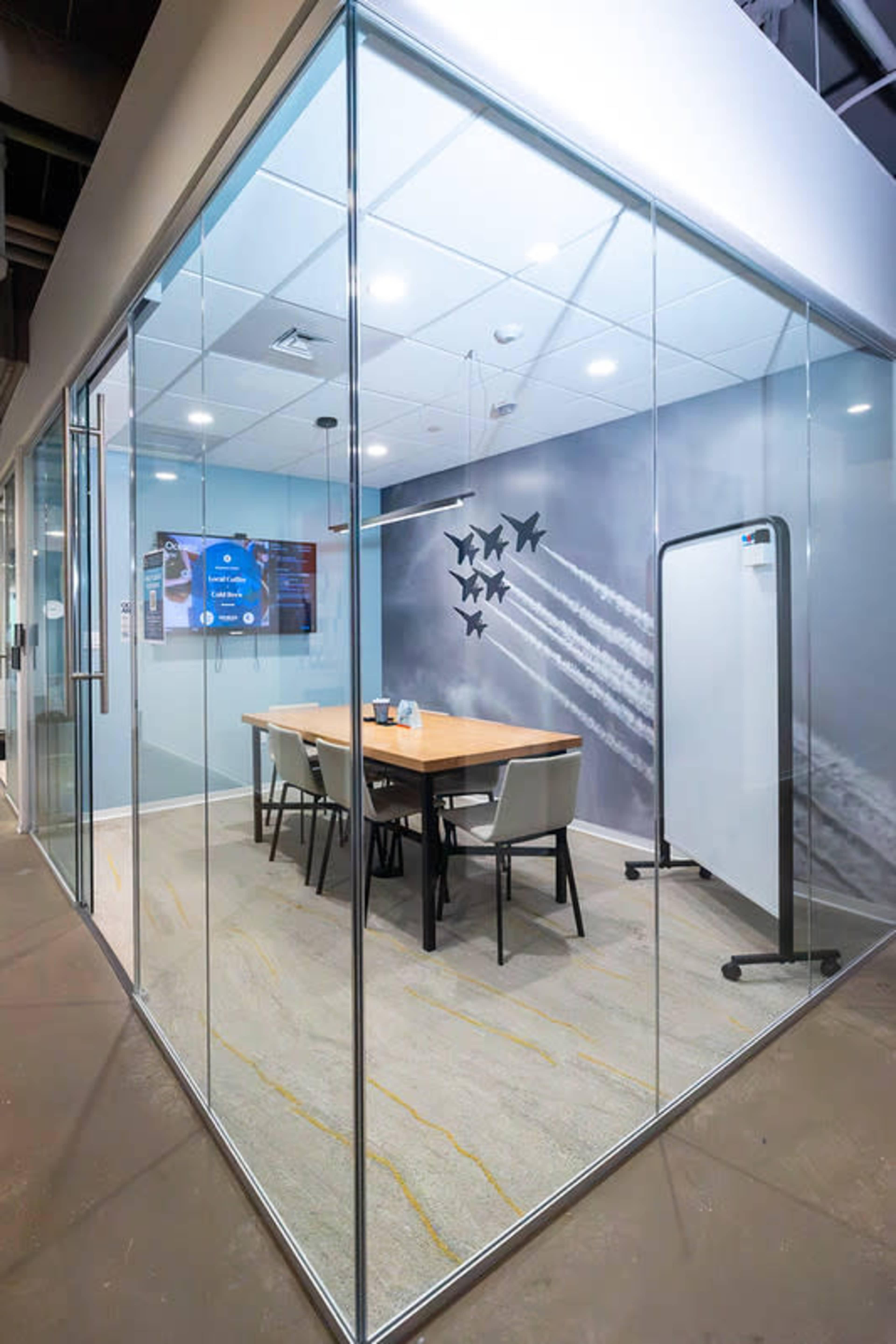 The image shows a modern conference room enclosed in glass, featuring a wooden table with chairs, a whiteboard, and a wall displaying a video screen and decorative artwork of planes.