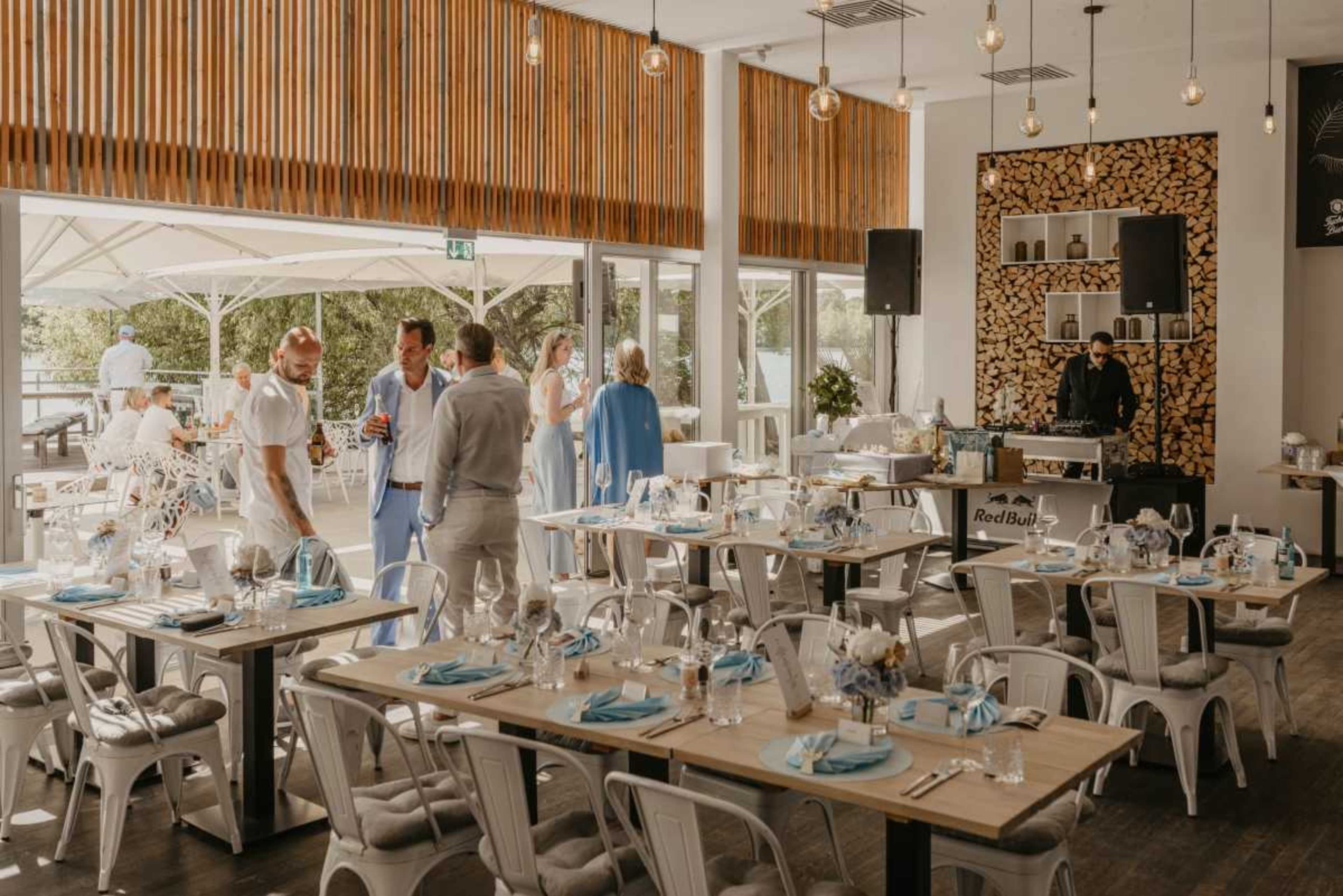 A bright and modern restaurant features tables set with blue accents, while guests mingle and a DJ plays music in the corner.