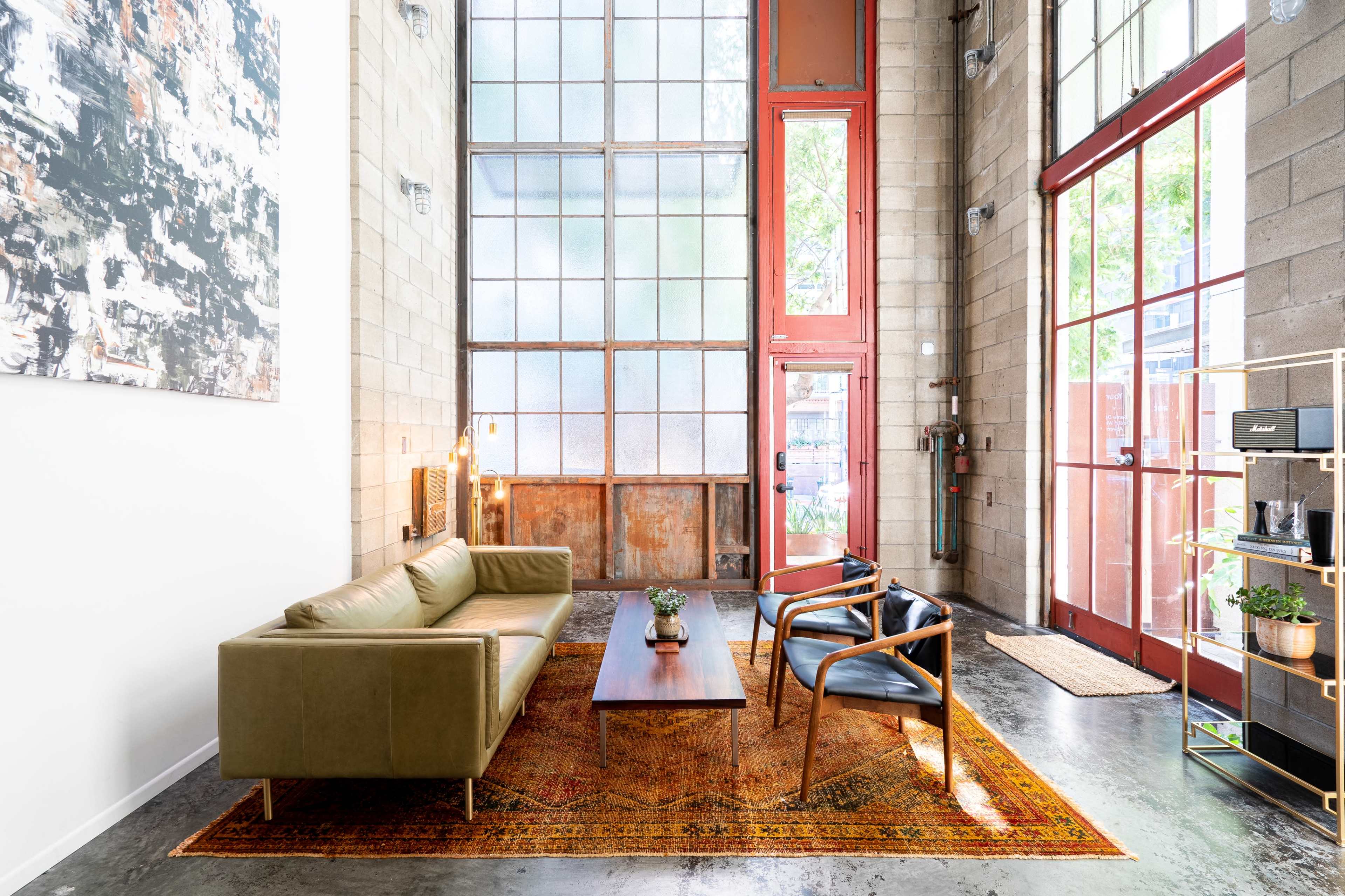 The image shows a modern living room with a green leather sofa, two black chairs, a wooden coffee table, and large windows allowing natural light to enter.