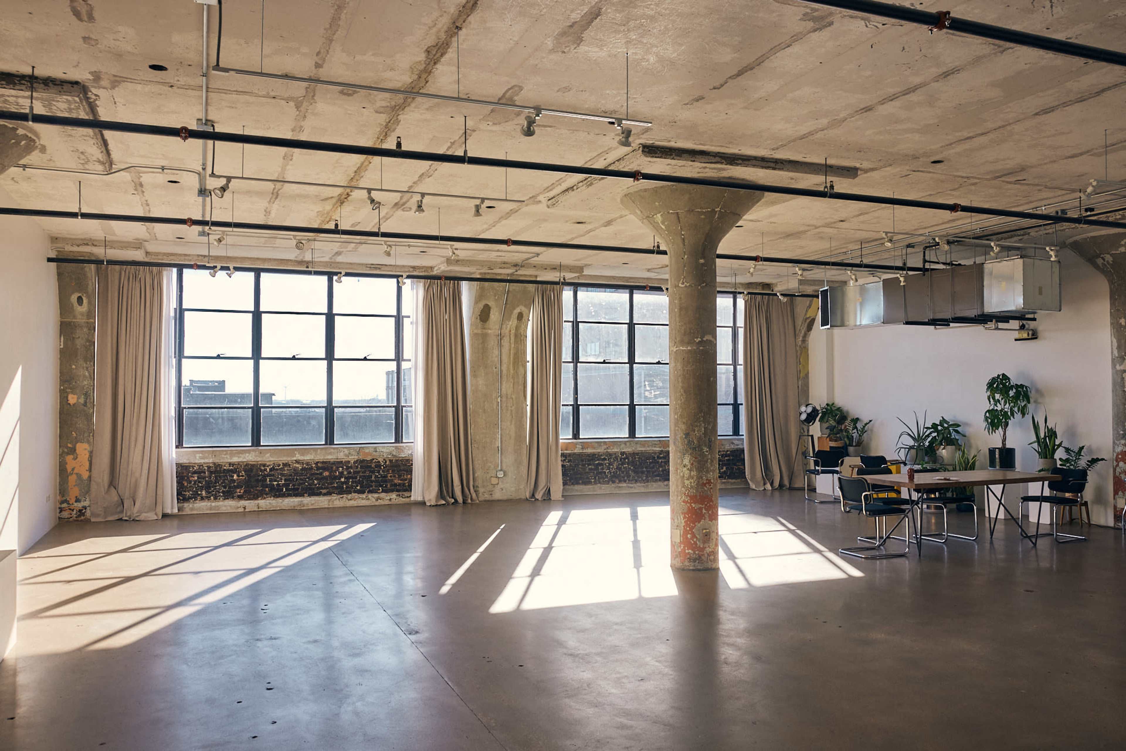 The image shows a spacious industrial-style room with large windows, concrete columns, and minimal furnishings, illuminated by natural light.