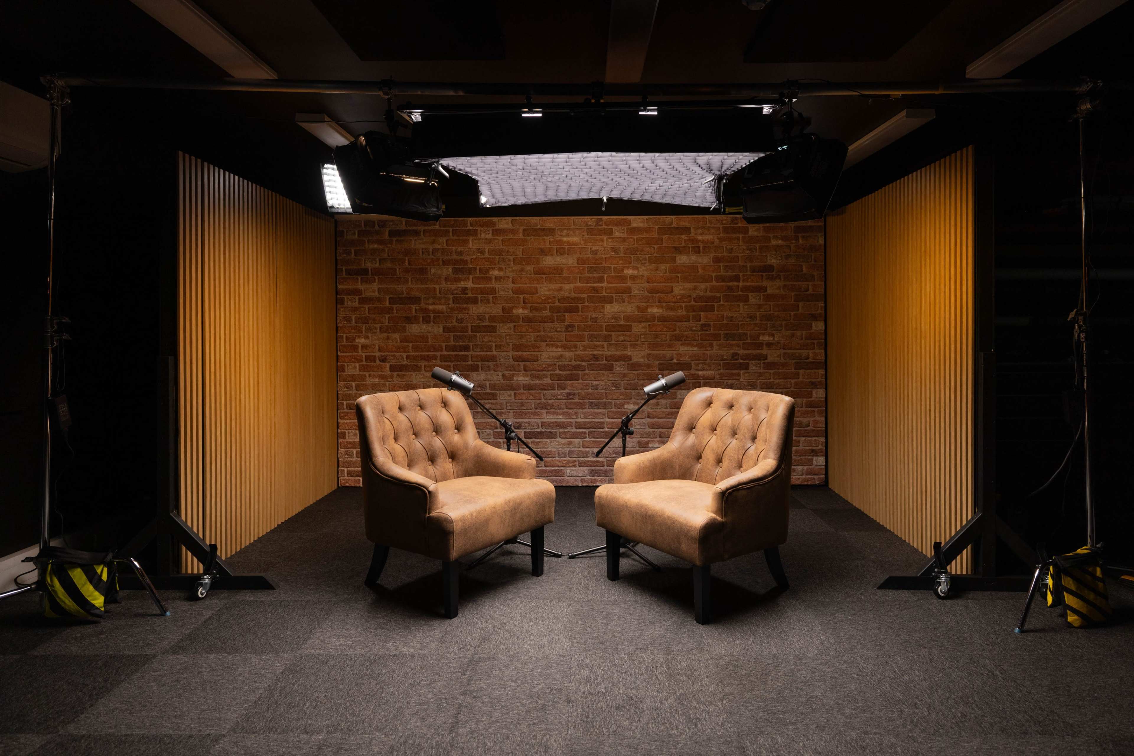 The image shows two brown upholstered chairs facing each other, with microphones positioned beside them, in a studio setting featuring a brick wall and wooden panels.
