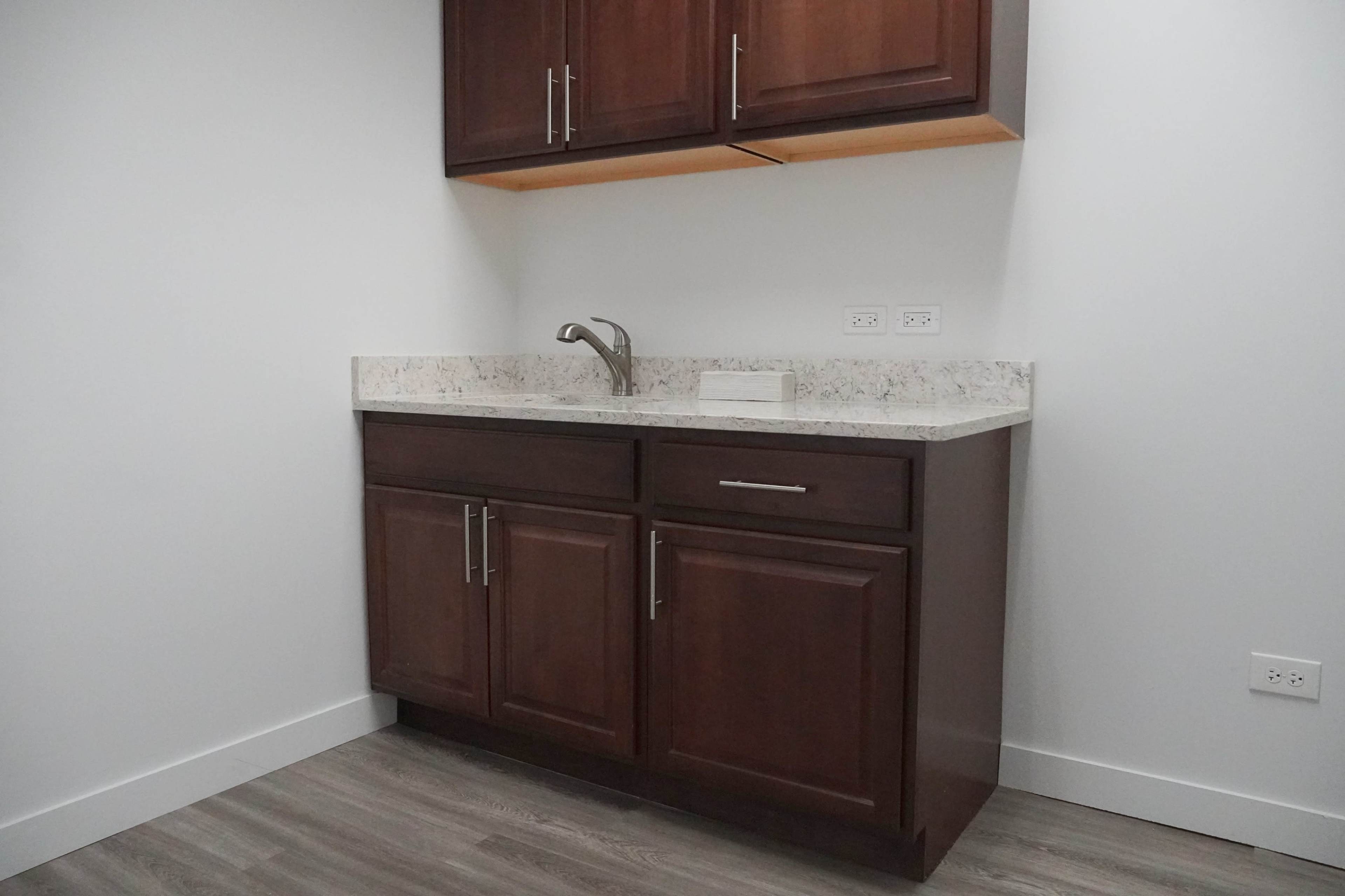 A corner kitchen area features dark wooden cabinets above and below a light-colored countertop with a sink and faucet.