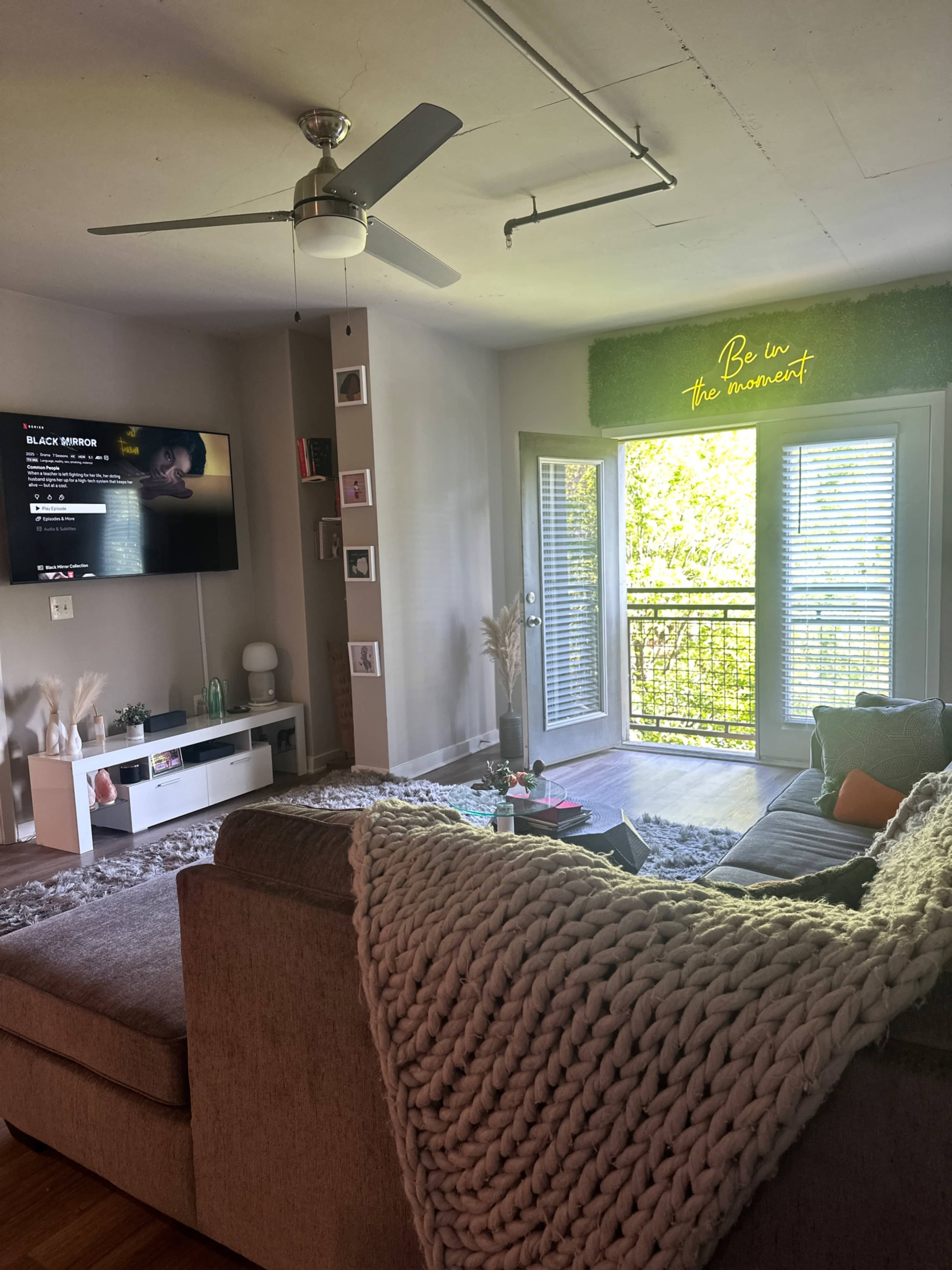 The living room features a sectional couch with a large blanket, a television mounted on the wall displaying a show, and a balcony with open glass doors letting in natural light.