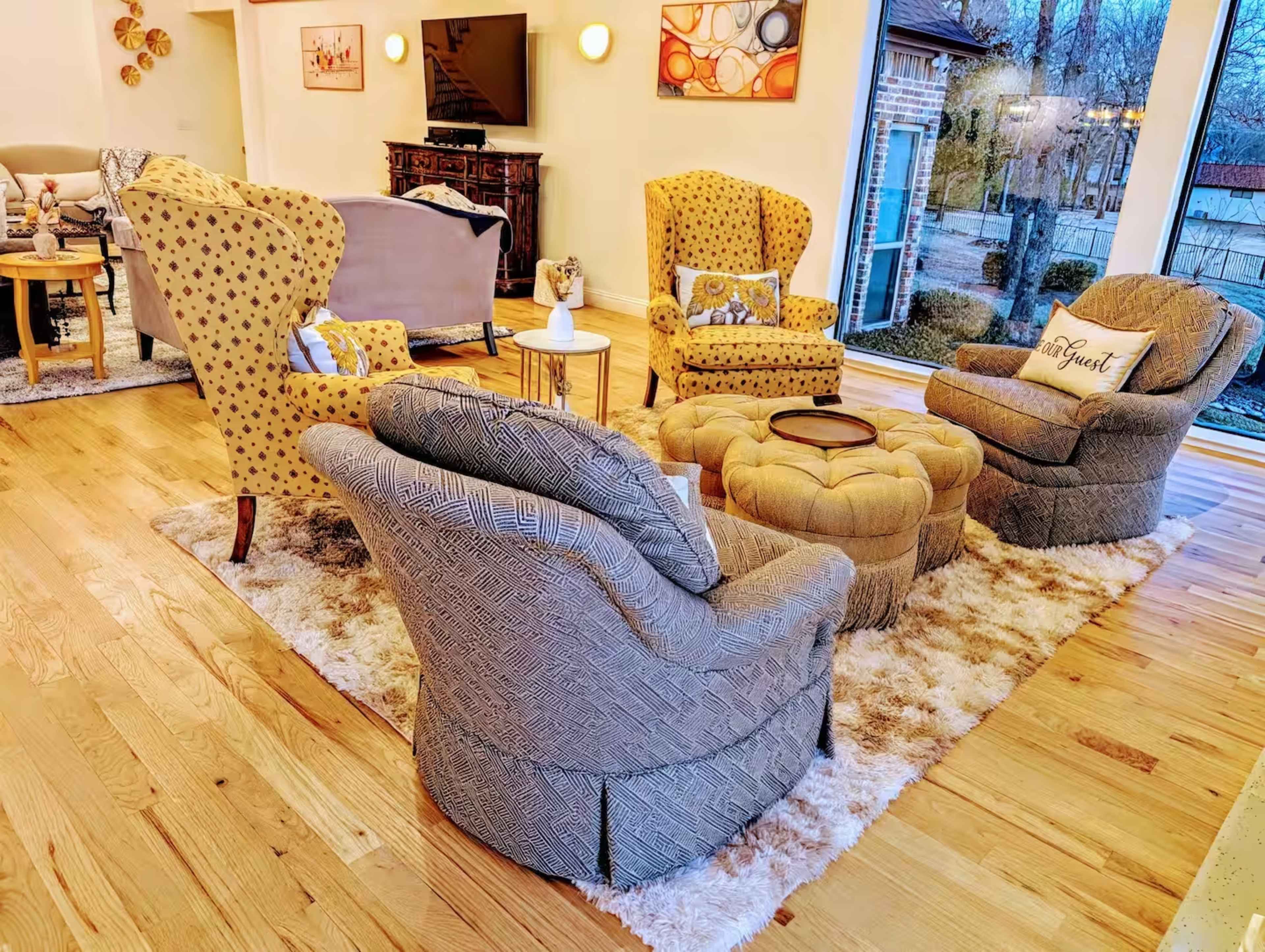 The living room features two patterned yellow chairs and a gray upholstered chair arranged around a round tufted coffee table on a rug.