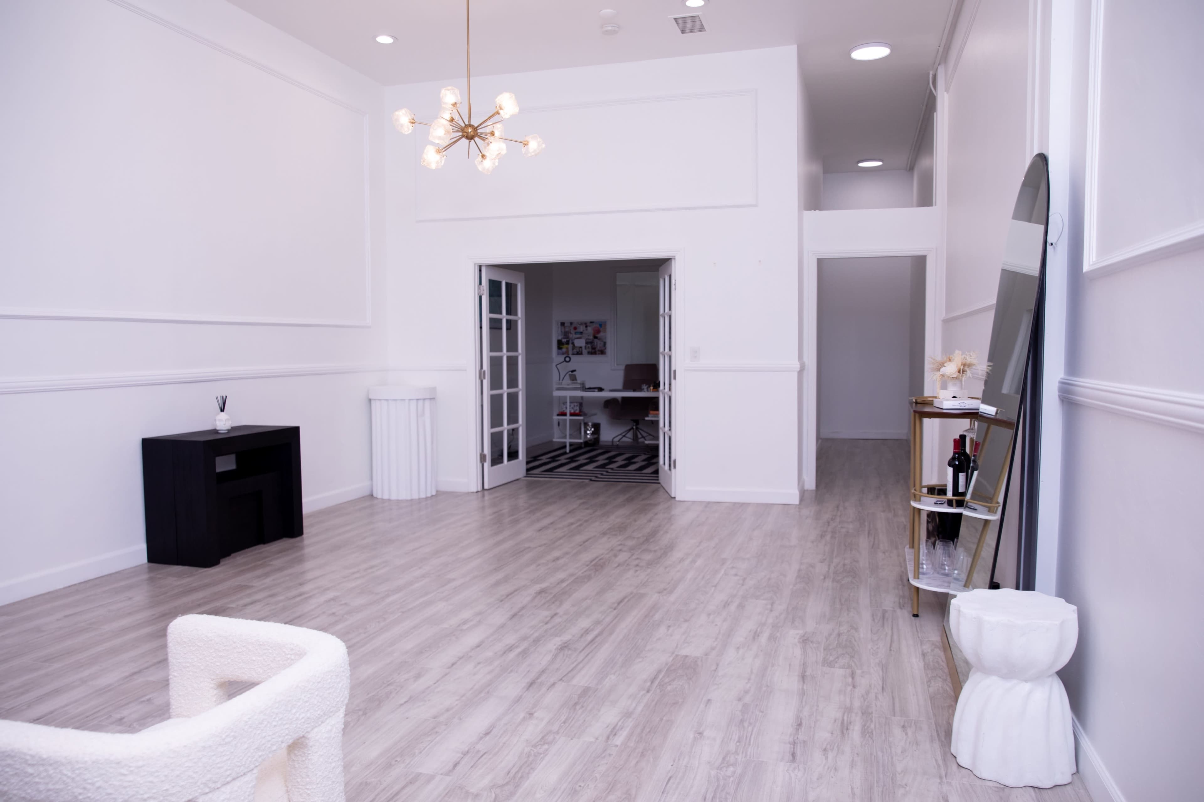 A spacious, well-lit room with wooden flooring, featuring a black furniture piece, a white waste bin, and an entrance to another room in the background.